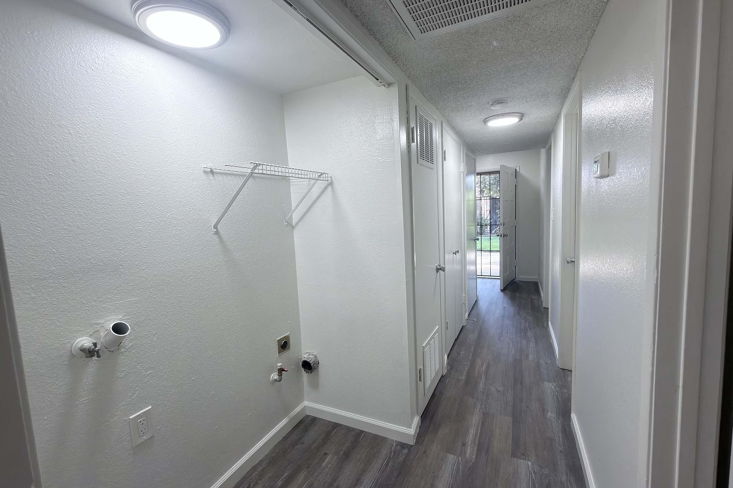A well-lit hallway featuring white walls and a wooden floor. On one side, there is a wire shelf mounted on the wall, while plumbing fixtures are visible below. Doors lead to adjacent rooms, with natural light coming from an open door at the end of the hallway, providing a welcoming atmosphere.