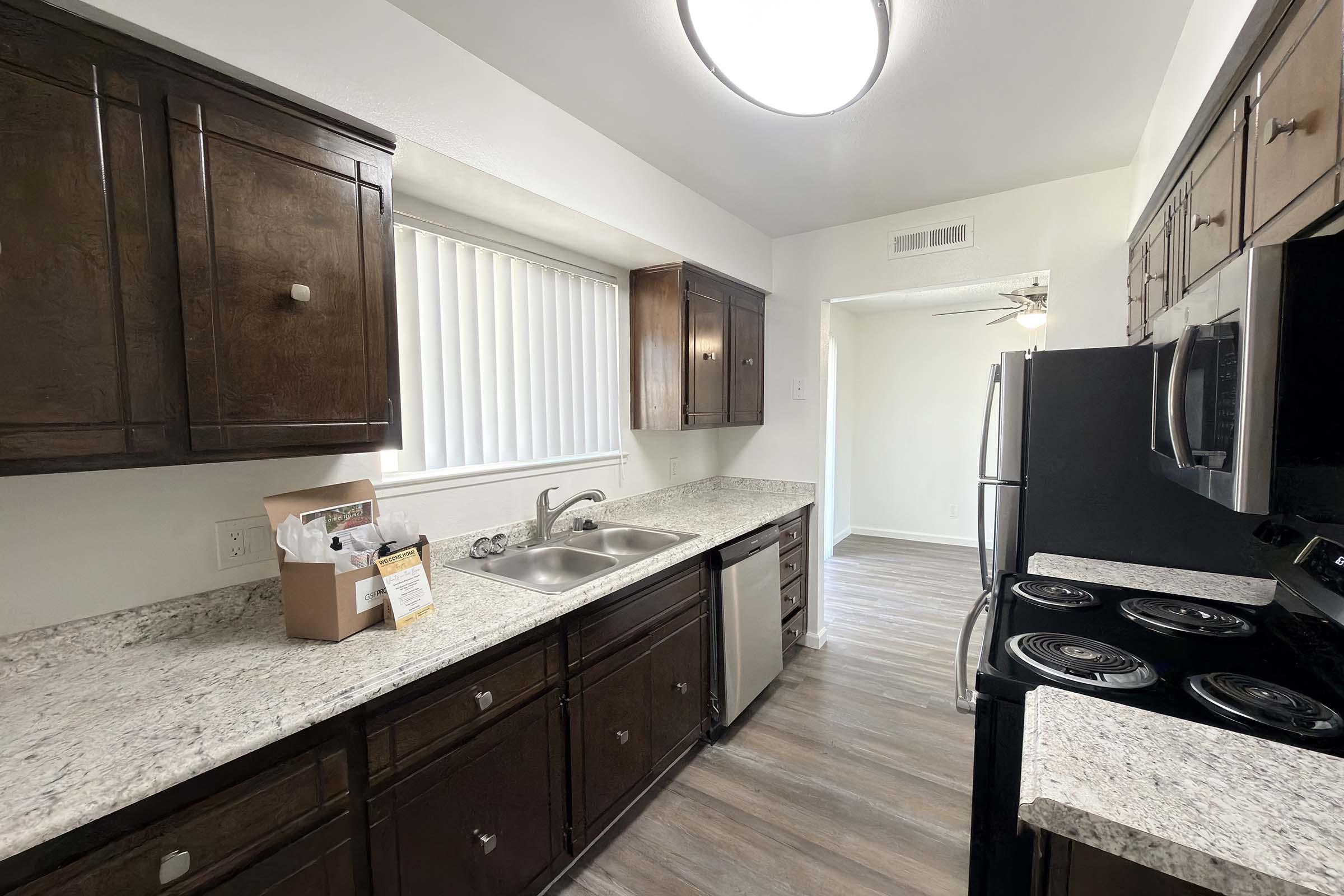 A modern kitchen featuring dark wood cabinets, a double sink with a window above, and granite countertops. The area includes stainless steel appliances, including a refrigerator and stove. A door at the end leads to another room, and the floor is finished with light-colored laminate. Bright overhead lighting enhances the space.