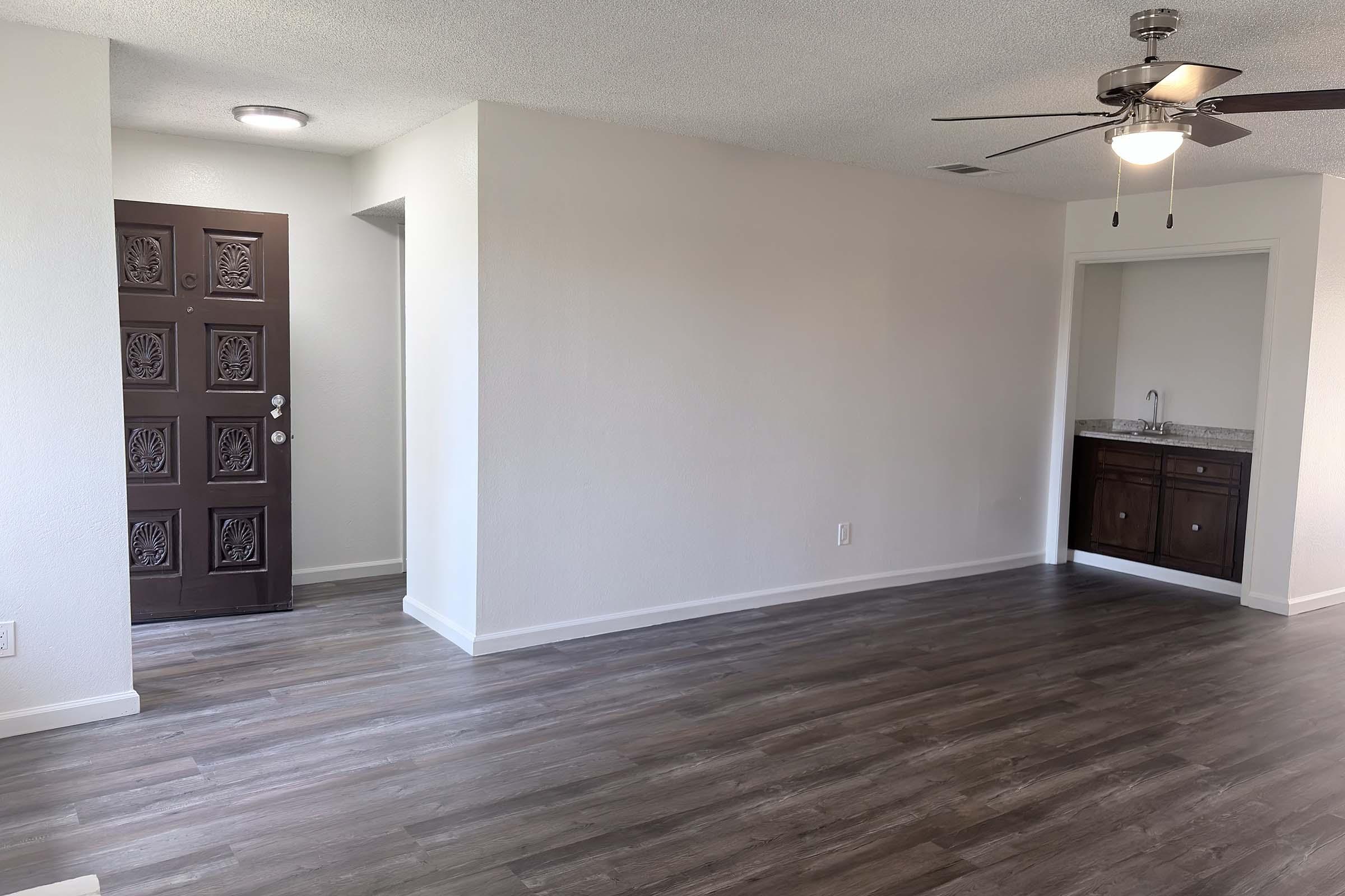 A well-lit living room with light gray walls and wooden flooring. There is a decorative front door on the left, leading to a hallway. In the corner, there's a small kitchenette area with a dark wood cabinet. A ceiling fan is visible, adding to the modern aesthetic of the space.