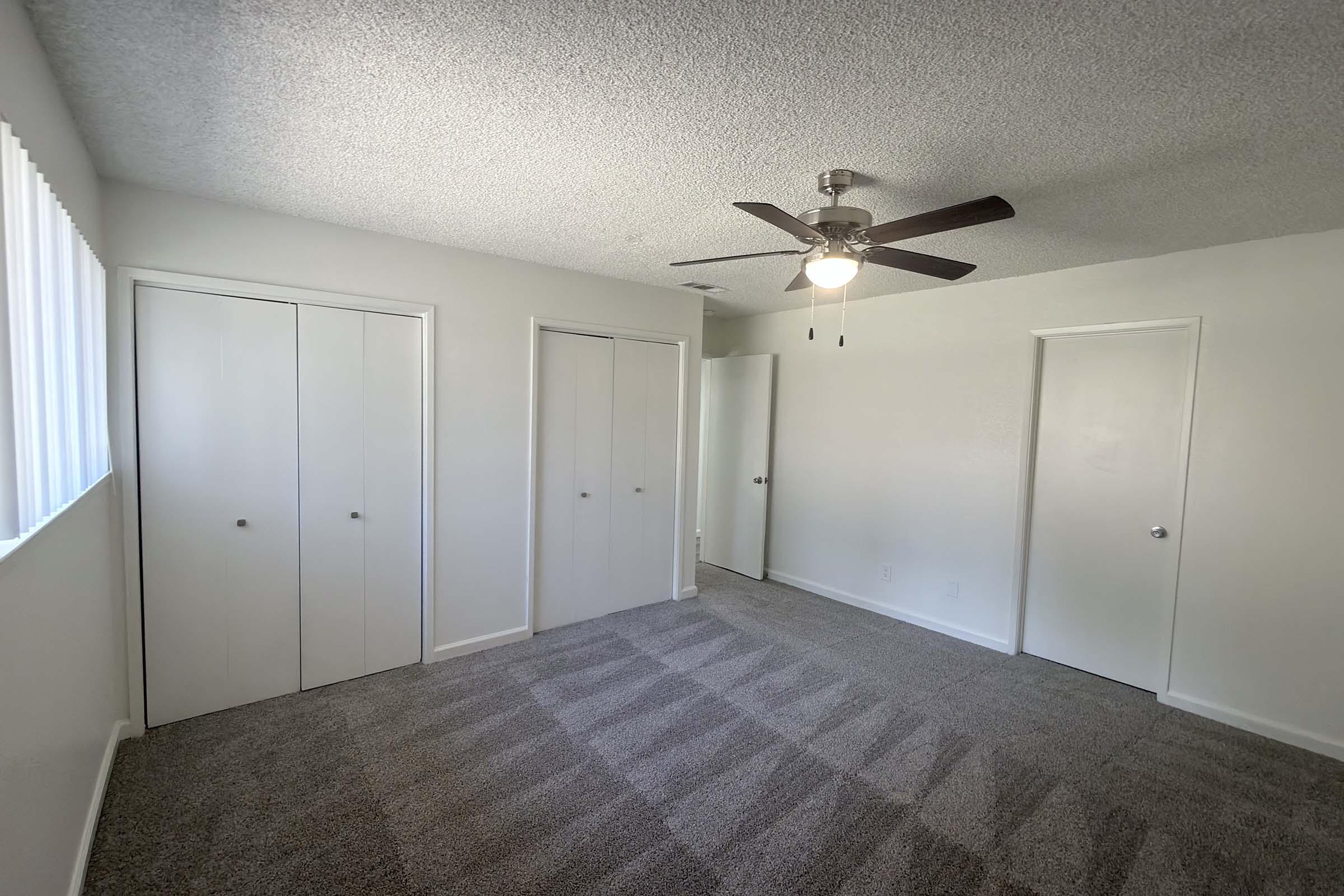 A spacious room featuring light gray carpet, two sets of white double-door closets, and a ceiling fan. There are two doors leading to other areas, and a window with blinds allowing natural light to enter. The walls are painted a light color, creating a bright, open atmosphere.
