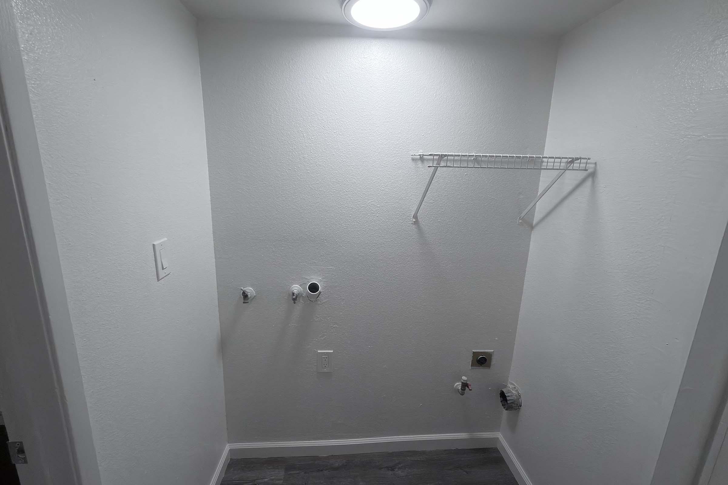 A clean and empty laundry room with white walls. There are plumbing connections on the left wall for water supply and drainage. A small shelf is mounted on the right wall, and a ceiling light brightens the space. The floor features dark tiles, adding contrast to the room's bright walls.