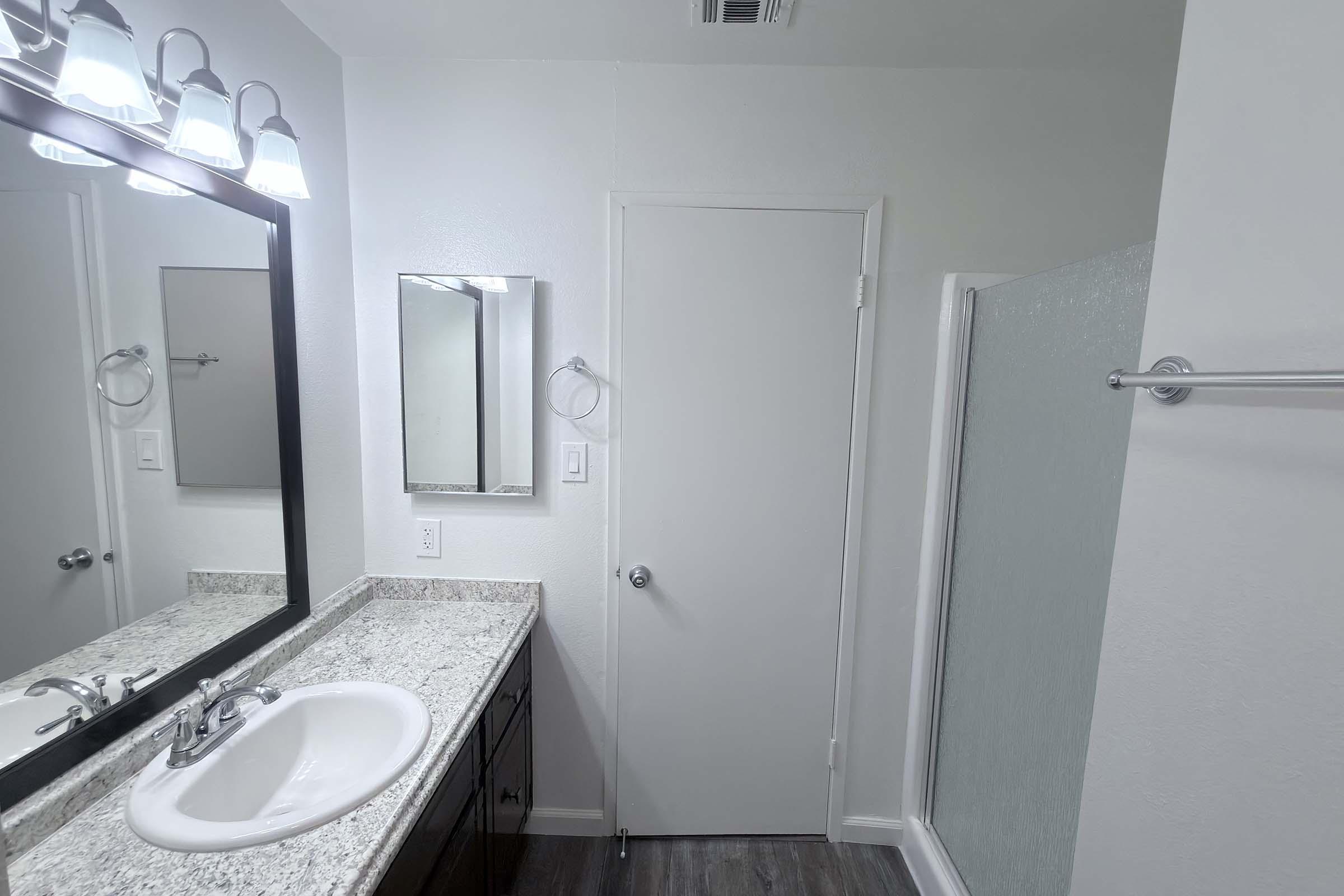 A modern bathroom featuring a granite countertop sink with a mirror above, a closed door, and a frosted glass shower enclosure. Soft lighting from four ceiling lamps enhances the bright, clean atmosphere, with neutral-colored walls and a wood-style floor completing the design.