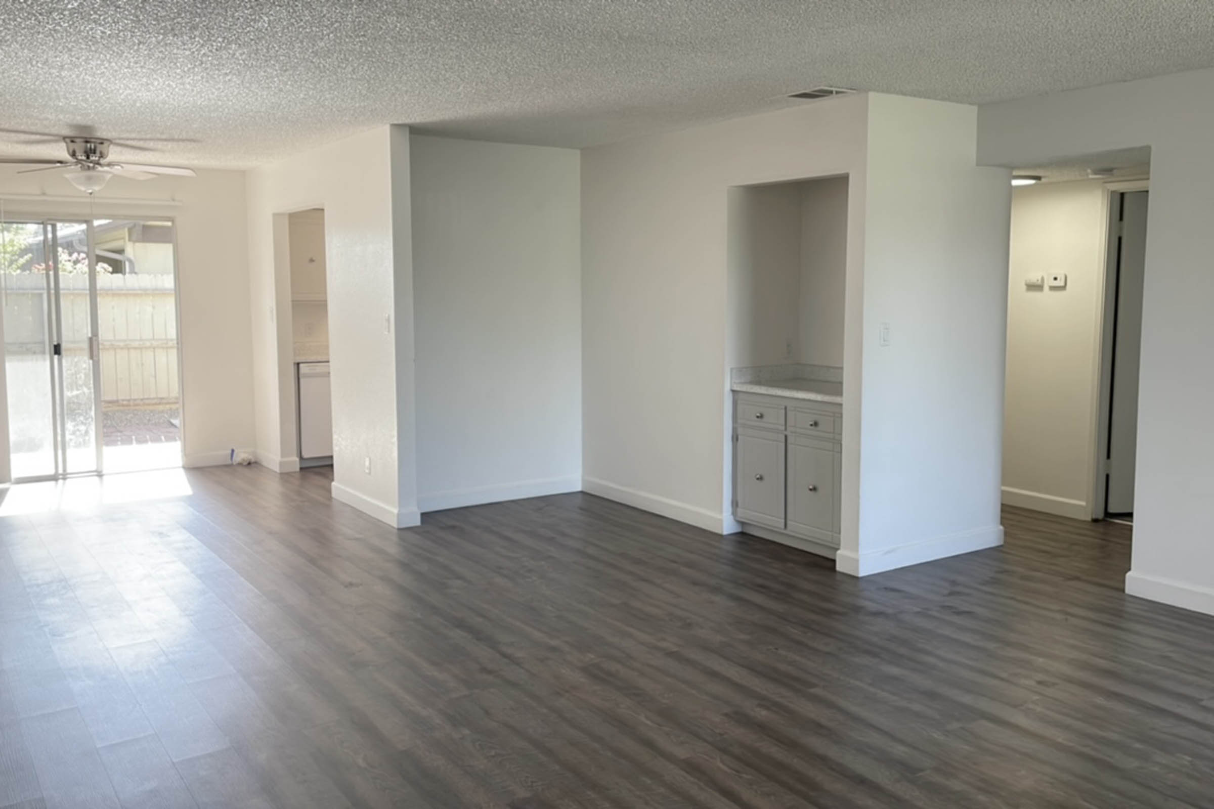Spacious, well-lit room with neutral-colored walls and laminate flooring. Features a ceiling fan and an adjacent kitchenette area with a counter. Open layout leads to a sliding glass door bathed in natural light, opening to an outdoor space. Includes a small built-in cabinet unit.