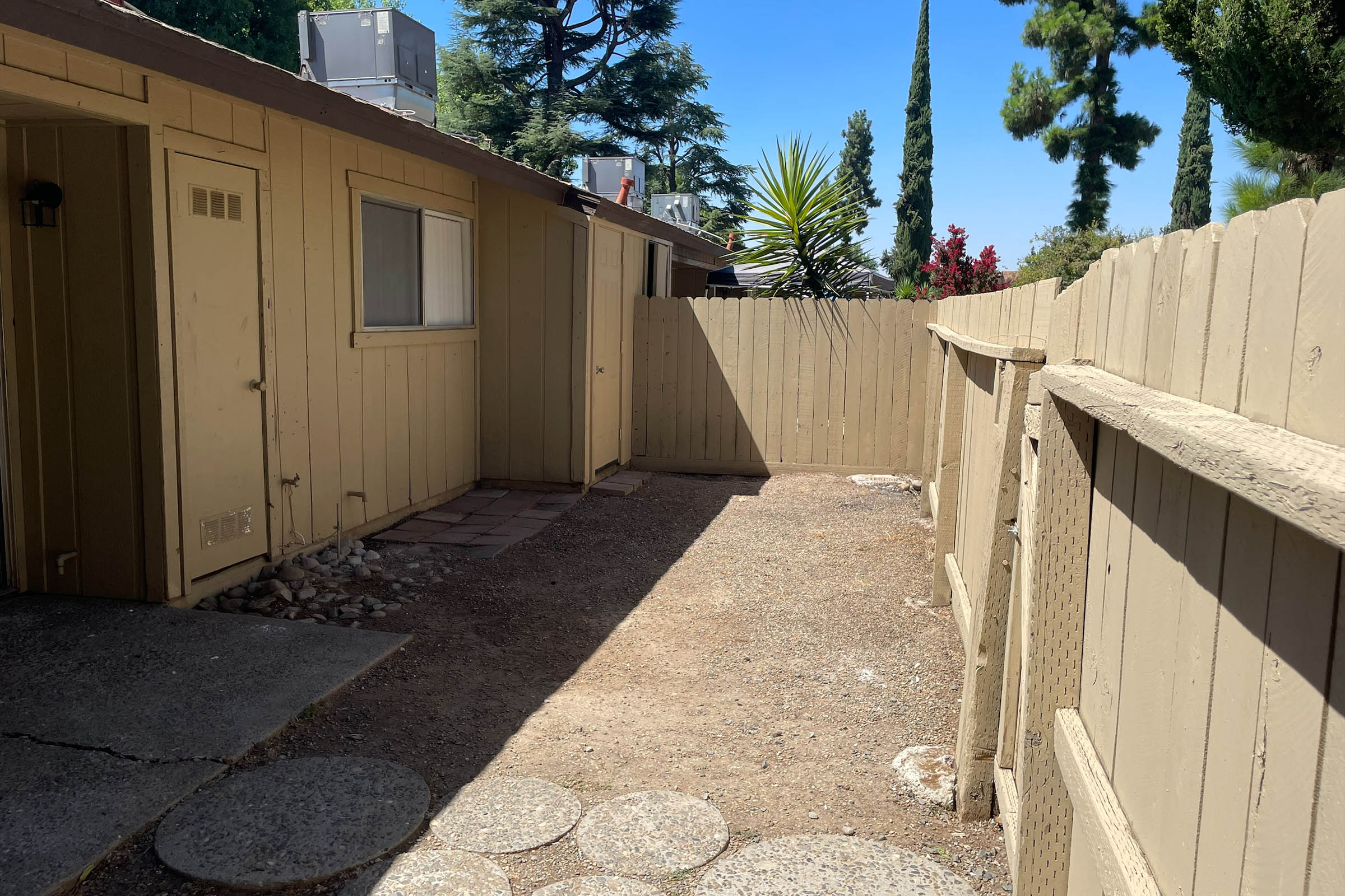 A narrow outdoor space between two buildings, featuring a wooden fence and a path made of stone slabs. The ground is covered with gravel, and there are a few plants visible, including tall trees in the background. The sky is clear and blue, indicating a bright day.