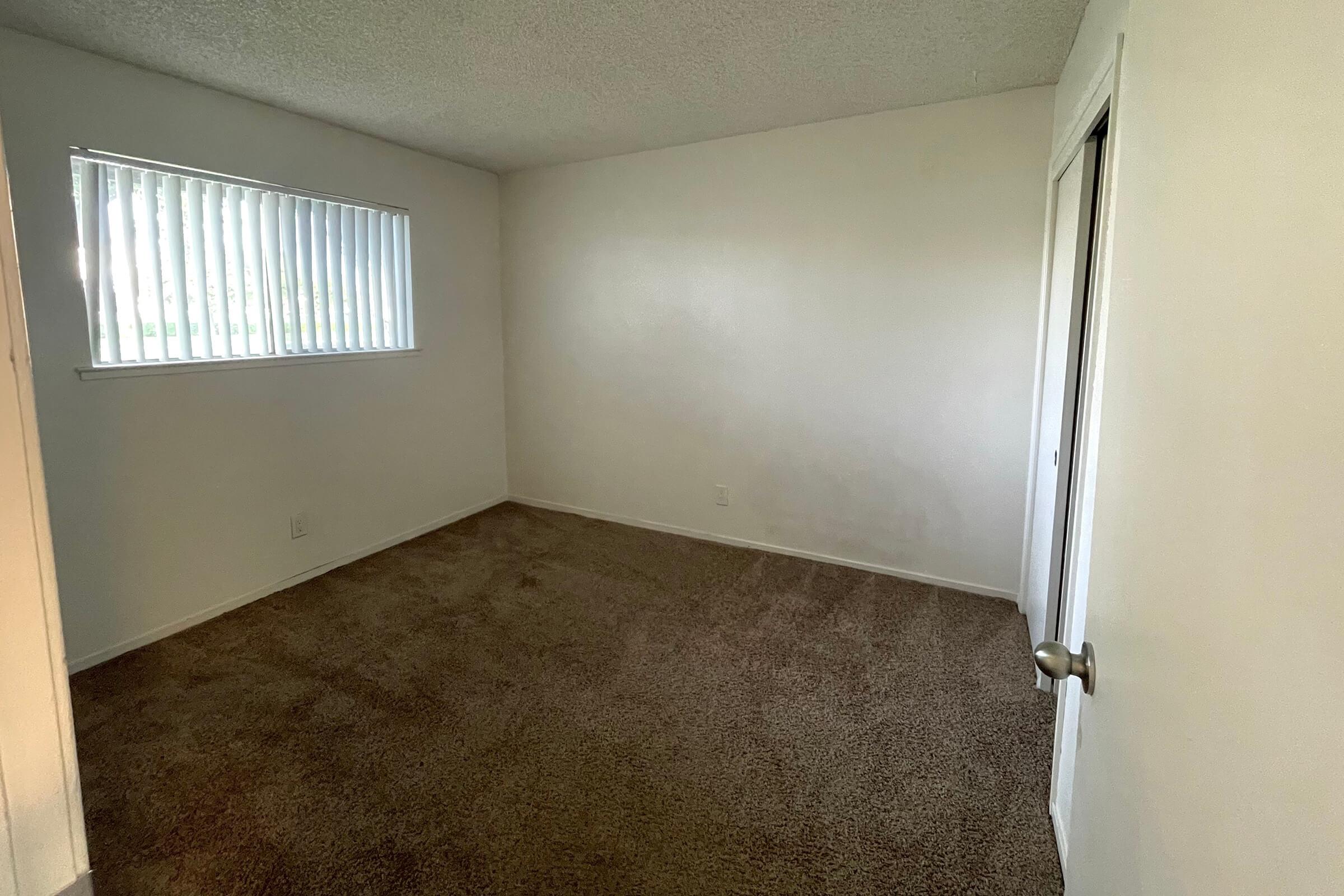 A vacant room with beige carpet and white walls. There's a window with vertical blinds on the left side, allowing natural light to enter. A closed door is visible on the right side of the image. The room appears clean and unfurnished, creating a spacious and minimalist atmosphere.