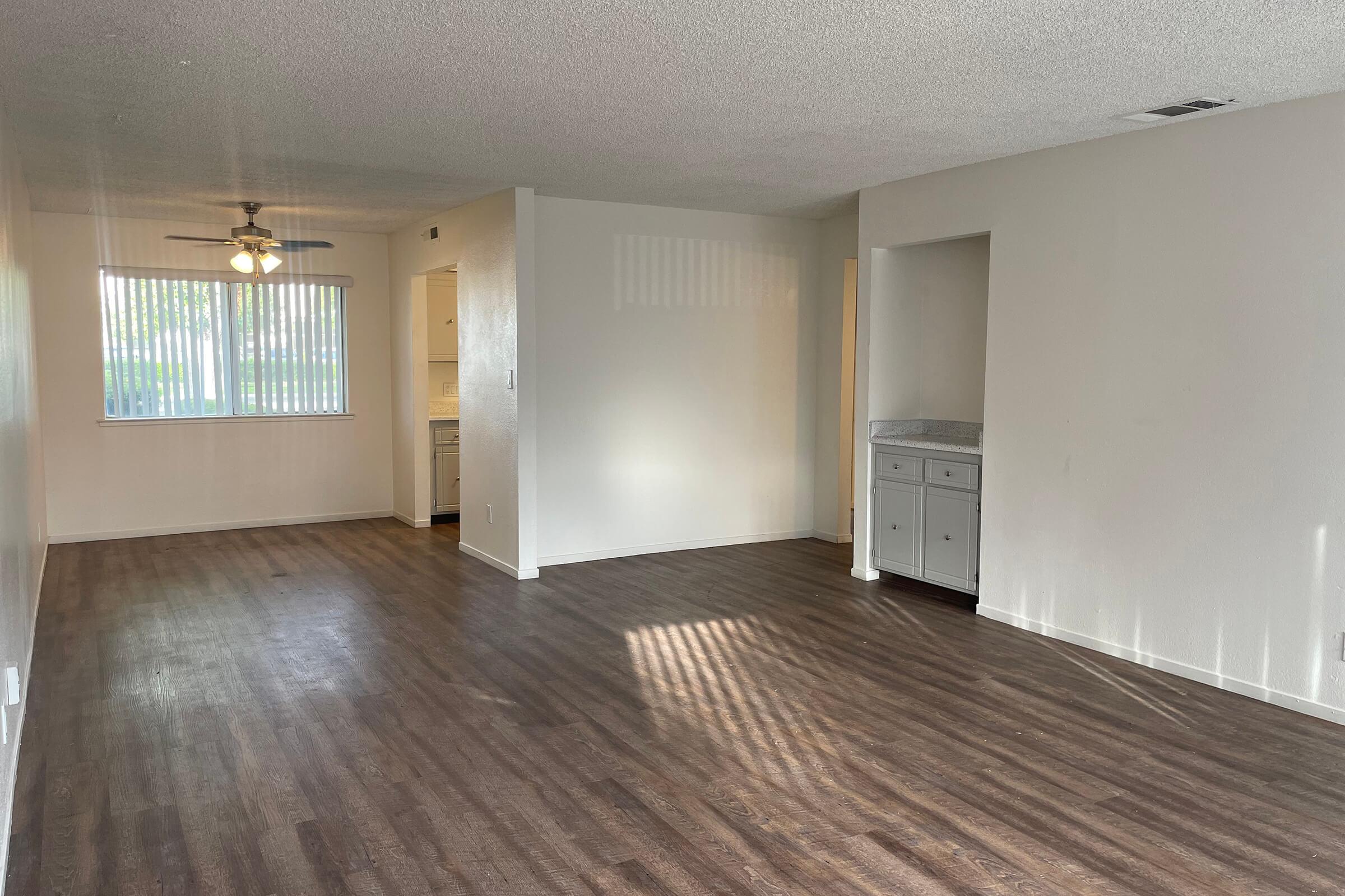A spacious, empty living area with light-colored walls and wooden flooring. A ceiling fan hangs from the ceiling. On one side, there is a window with vertical blinds allowing natural light to enter. In the background, a small kitchenette can be seen, with cabinetry and a countertop, creating an open-plan feel.