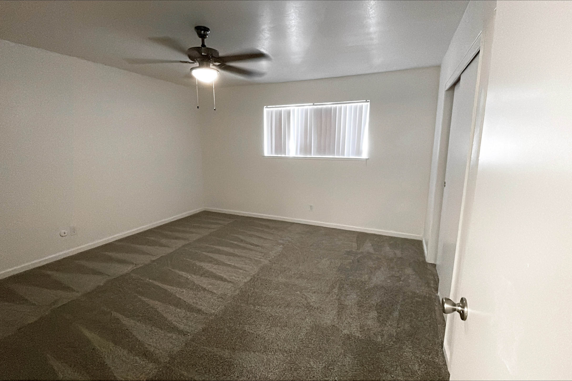 A spacious, empty room with light gray carpet flooring, a ceiling fan, and a window covered with sheer white blinds. The walls are painted white, and there are two doors visible, one of which is partially open. The room appears well-lit and ready for furnishing.