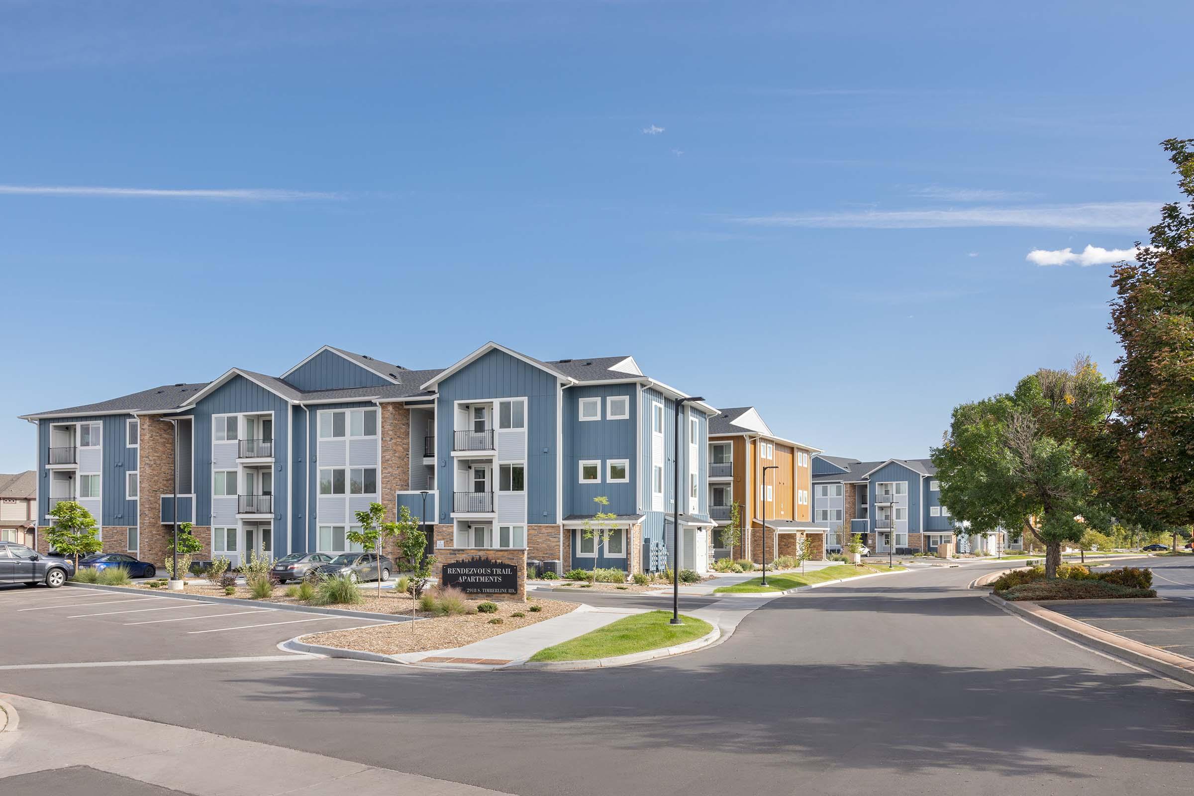 Modern apartment buildings in a suburban setting, featuring a mix of blue and orange facades. Well-maintained landscaping with trees, shrubs, and a neatly paved road. The sky is clear with a few clouds, creating a bright and inviting atmosphere.
