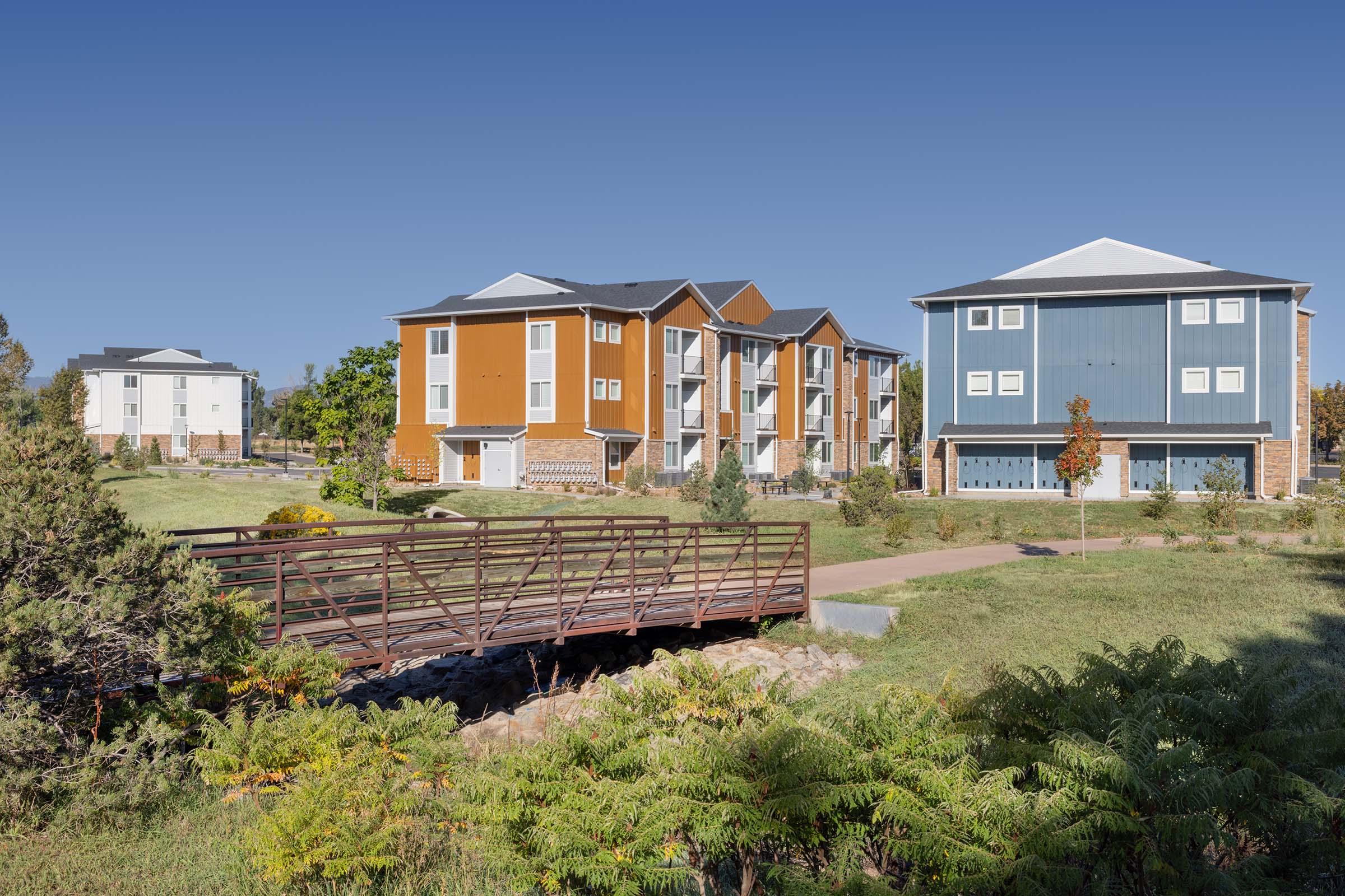 A modern residential complex featuring three multi-story buildings in varying colors, surrounded by well-kept landscaping. A small wooden bridge spans a path in the foreground, leading to green grass and trees in the area, under a clear blue sky.