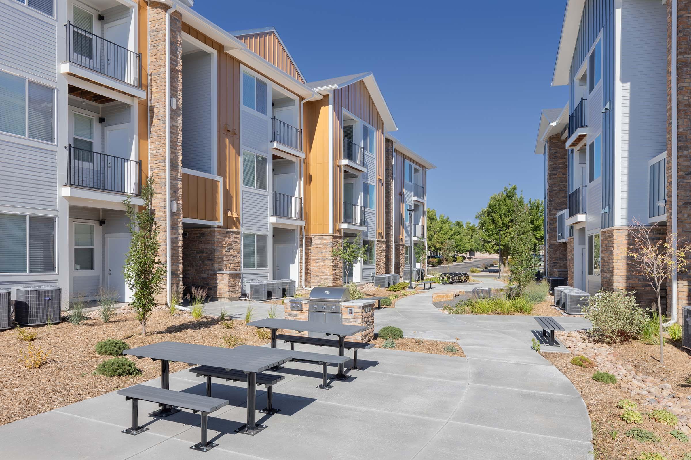A courtyard with two modern apartment buildings featuring a path between them. There is a picnic table and a grill area, surrounded by landscaped shrubs and trees, under a clear blue sky. The buildings have a mix of siding materials and balconies.