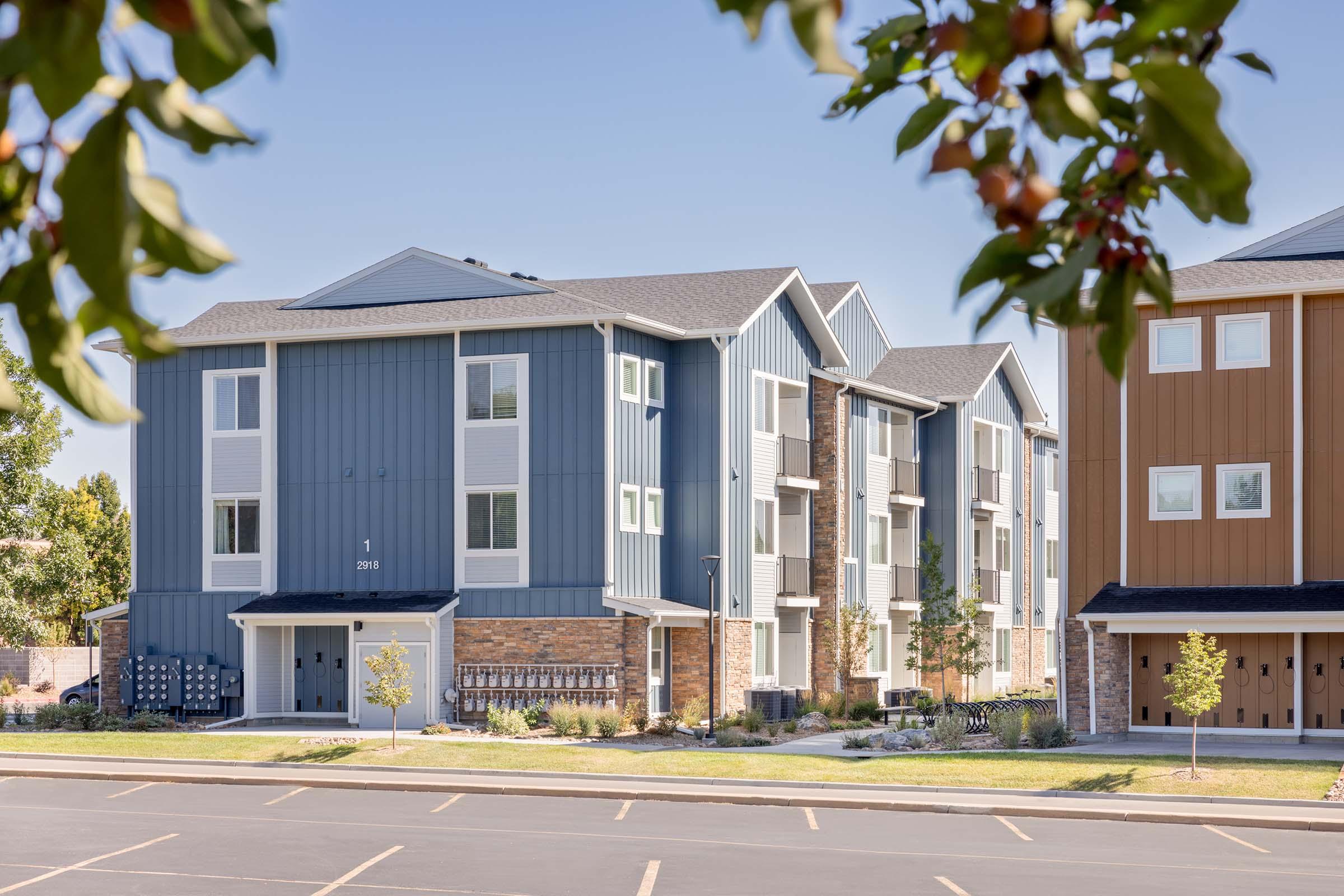 A view of two modern apartment buildings in a residential area. One building features a blue exterior with stone accents, while the other has a brown facade. The surroundings include small trees, manicured lawns, and a parking lot in front. The scene is bathed in natural light under a clear blue sky.