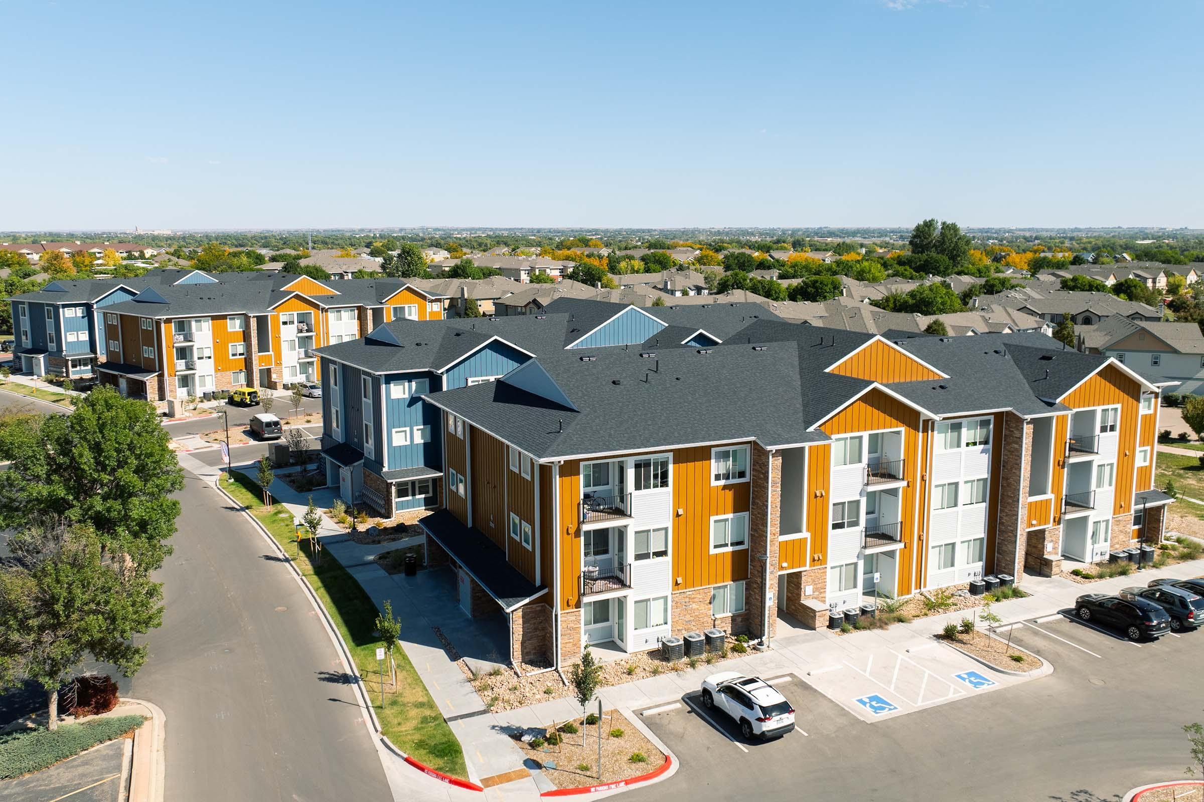 Aerial view of a modern residential complex with several multi-story buildings featuring colorful facades in orange, blue, and gray. The surrounding area includes well-maintained landscaping and parked cars. The sky is clear, and there are trees in the background, showcasing a suburban setting.