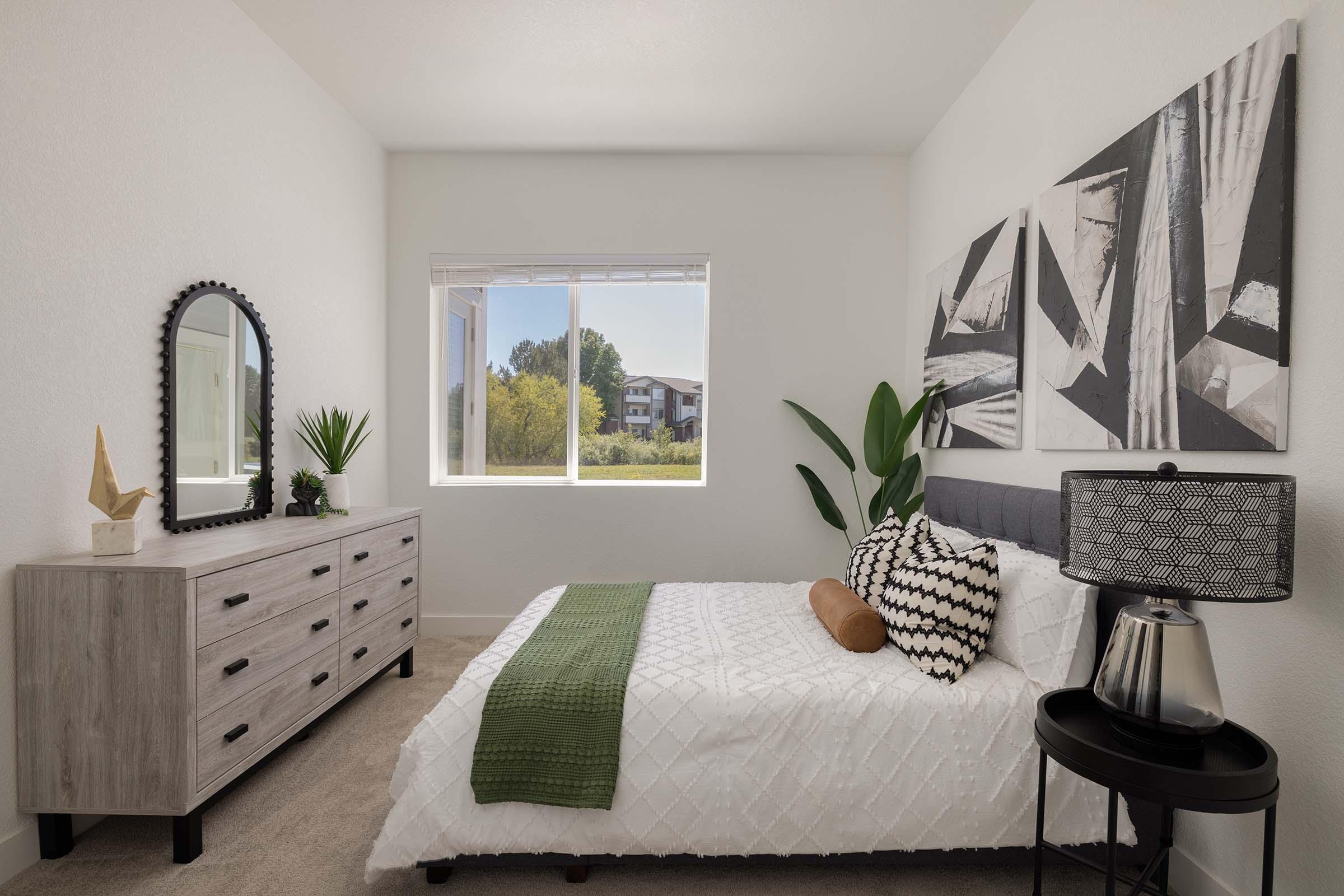 A cozy bedroom with a light color scheme. It features a neatly made bed with patterned pillows and a decorative throw. There's a stylish gray dresser with a round mirror and a small plant next to it. A large window lets in natural light, showcasing greenery outside and modern artwork on the wall.