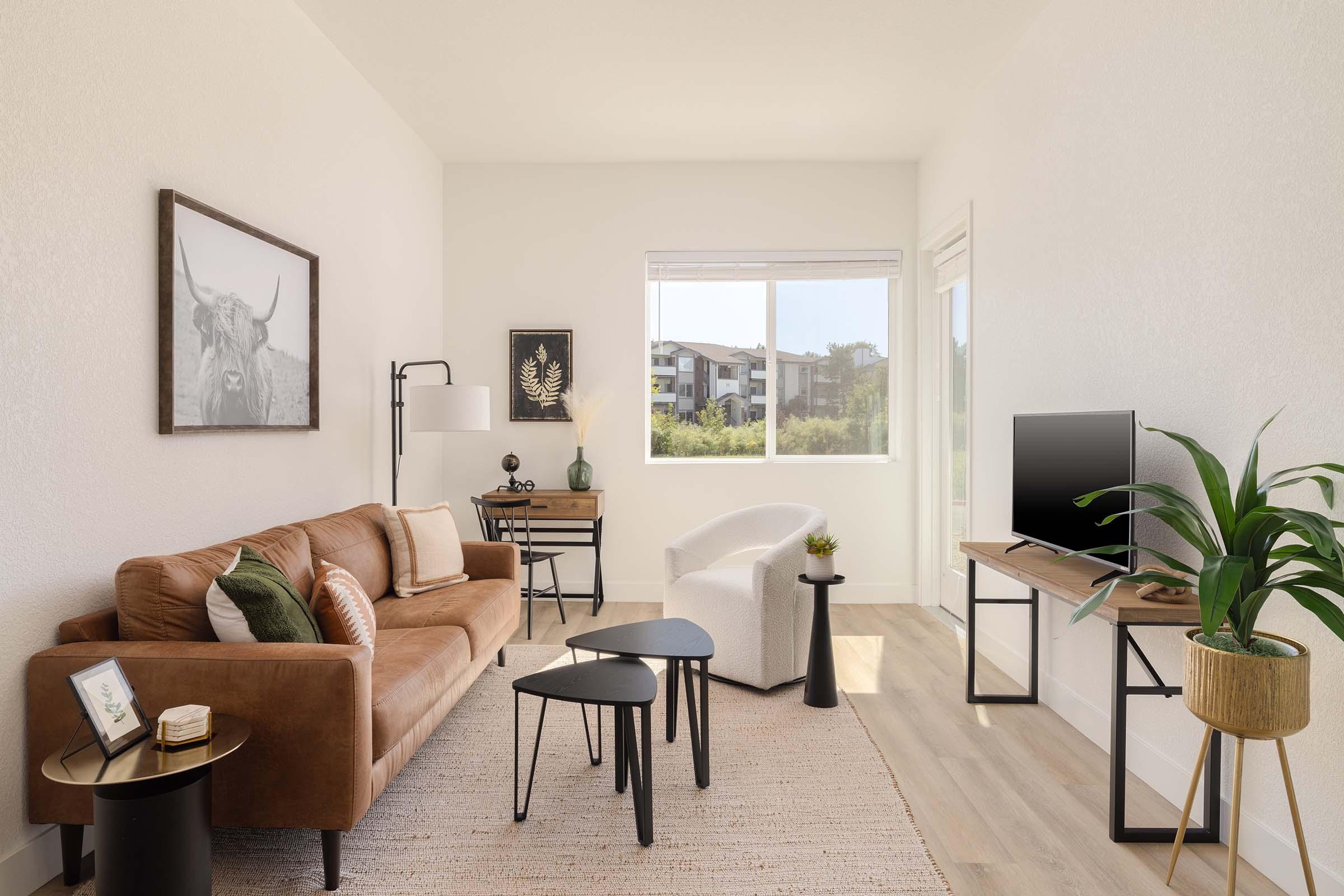 Interior of a modern living room featuring a brown leather sofa with cushions, a white armchair, and a stylish coffee table. A large window offers natural light, while a television sits on a wooden stand. Decorative elements include a framed picture of a cow, a plant, and a small side table with a decorative item.