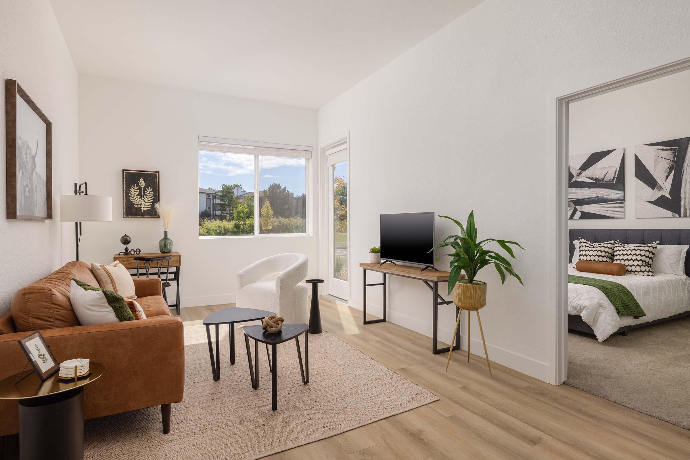 Cozy living room featuring a brown sofa, a small black table, and a light armchair. A flat-screen TV on a stand is visible, along with a decorative plant and framed artwork on the walls. A glimpse of a bedroom is seen through an open doorway, showcasing a bed with green and white bedding. Bright natural light illuminates the space.