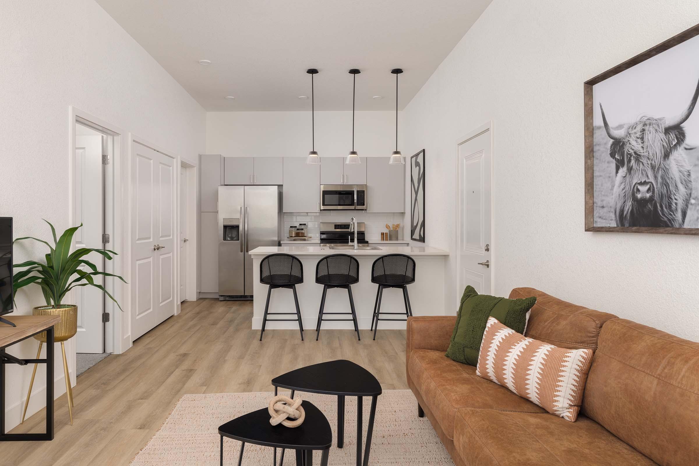 Modern living room and kitchen area featuring a brown sofa with decorative pillows, a black coffee table, and a plant. The kitchen has stainless steel appliances and three bar stools at a counter. There’s a large framed picture of a cow on the wall, with light wood flooring throughout.