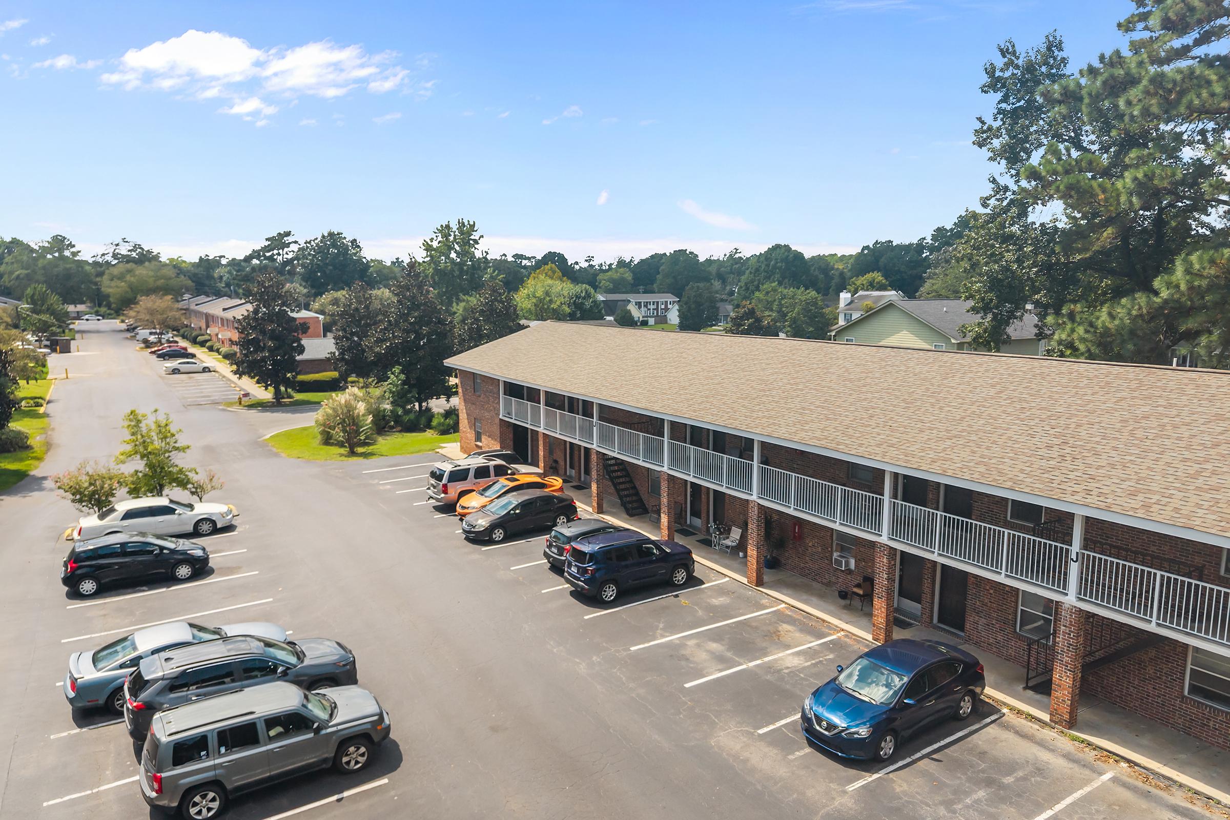 Aerial view of a residential apartment complex featuring two-story brick buildings. Several cars are parked in front of the apartments, with lush greenery surrounding the area. The sky is clear and blue, creating a bright and inviting atmosphere.