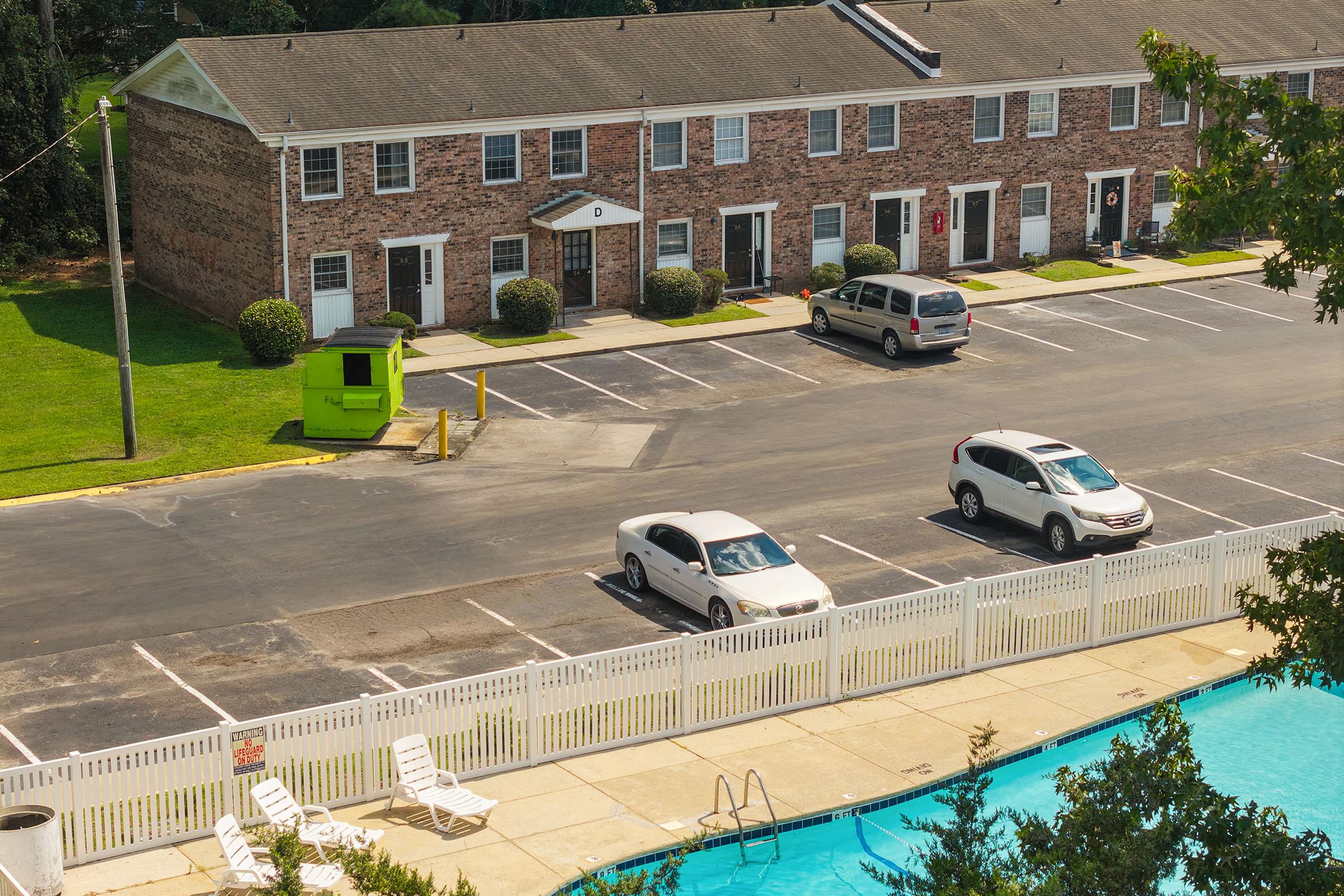 A view of a residential area featuring brick townhouses with white doors, parked cars in a lot, a green vending structure, and a swimming pool enclosed by a white fence. Lush greenery surrounds the area, creating a pleasant neighborhood atmosphere.