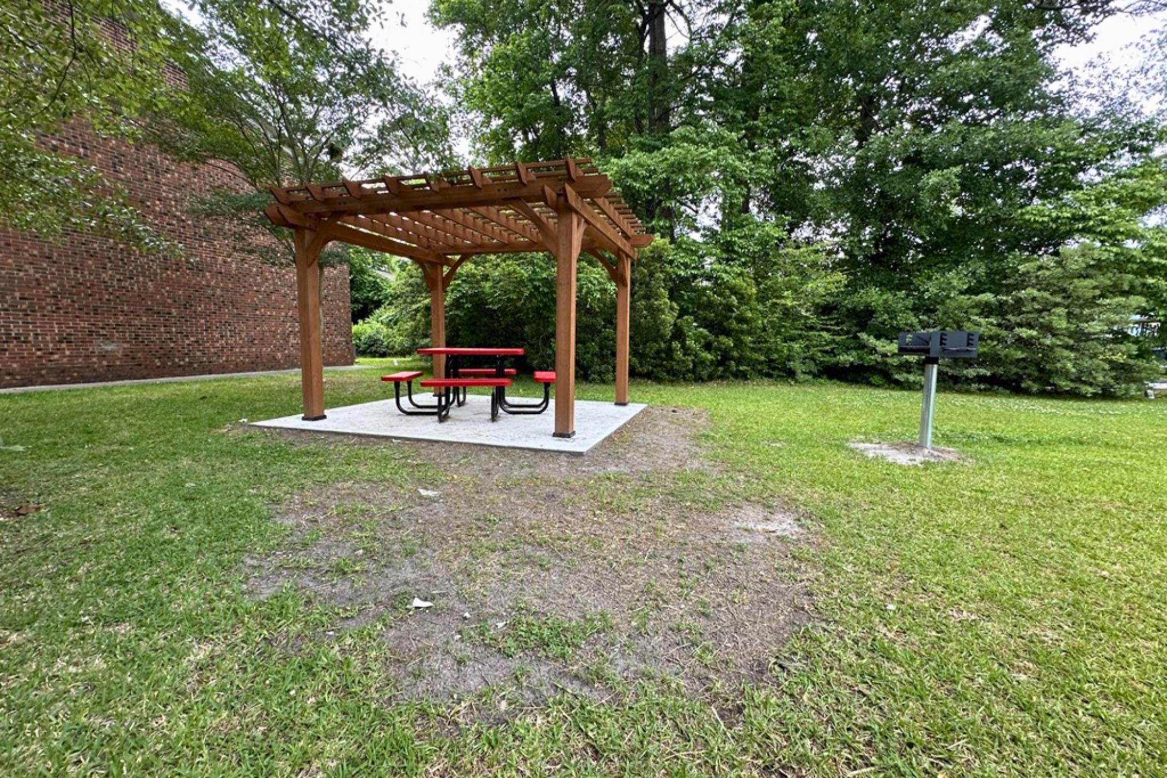 A small picnic area featuring a wooden pergola with a red picnic table and benches underneath, situated on grass. Nearby, there's a grill station, surrounded by trees and a brick building in the background.