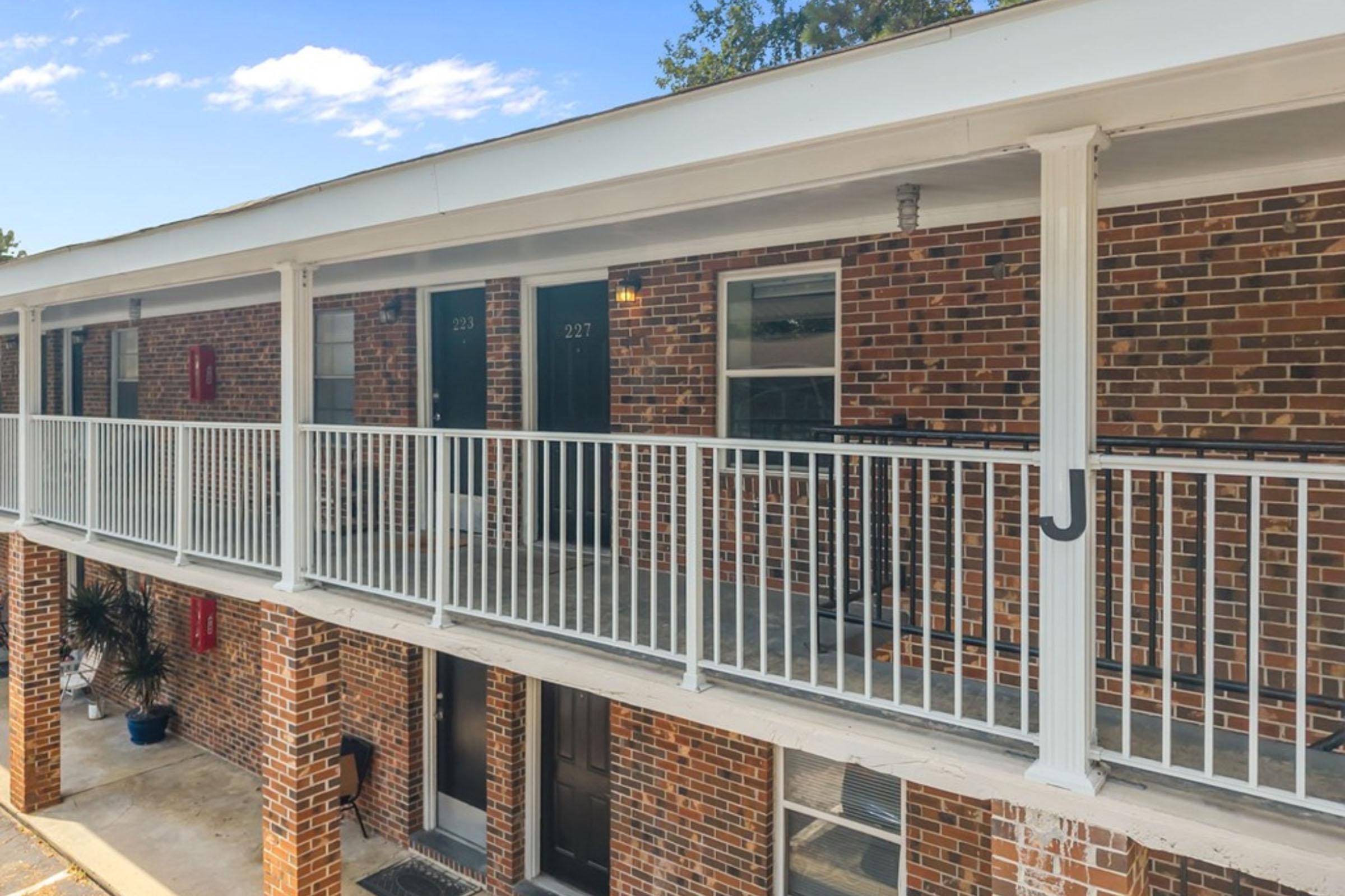 A two-story building featuring a row of apartment doors on a balcony, with brick walls and white railings. The doors are numbered 223 and 227. Potted plants are visible on the ground floor near the entrance, and the sky above is clear with a few clouds.