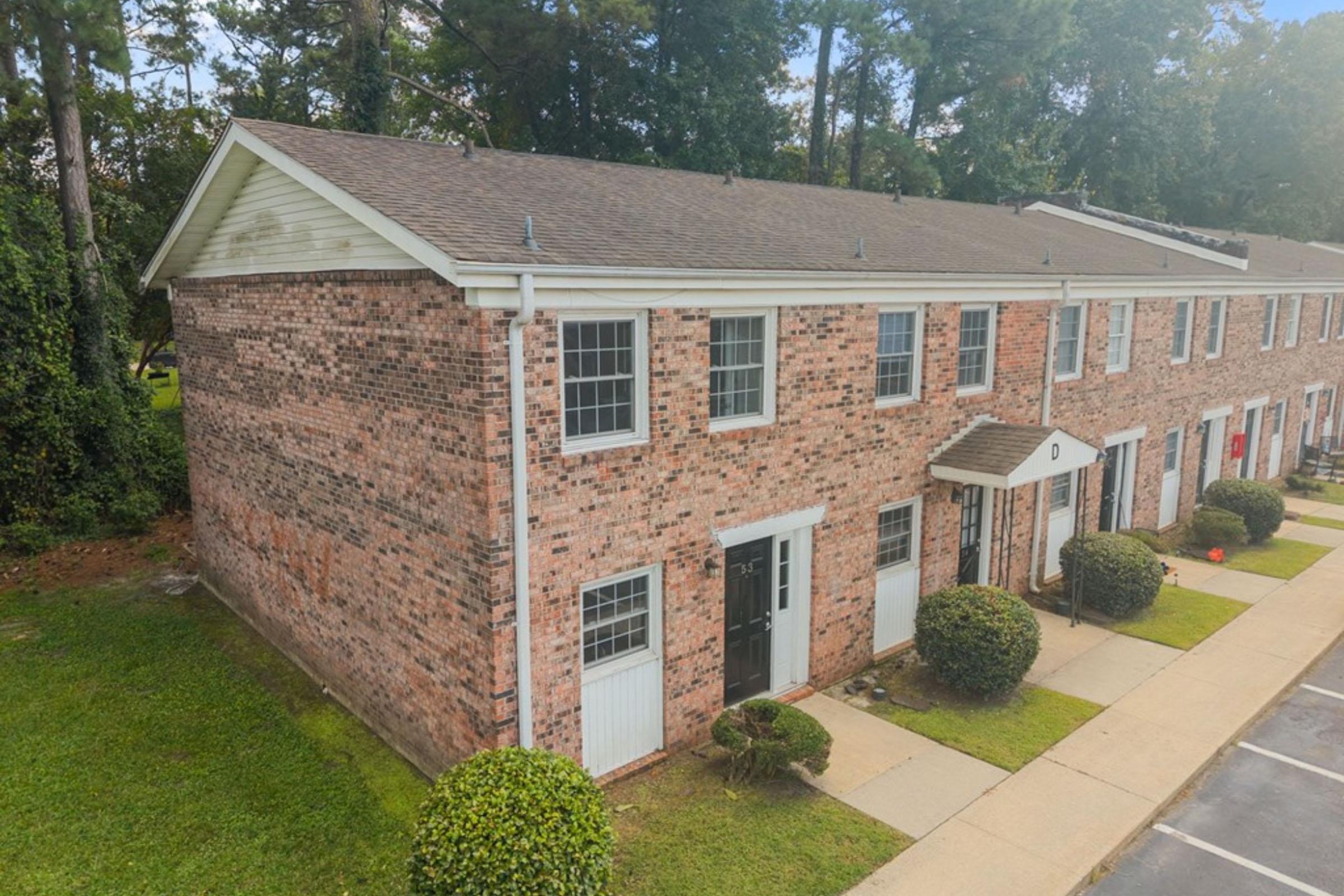 Aerial view of a brick townhouse complex featuring two-story buildings with multiple windows. The foreground shows a neatly paved sidewalk leading to the entrances, surrounded by landscaped areas with small shrubs and grass. Trees are visible in the background, creating a natural setting.
