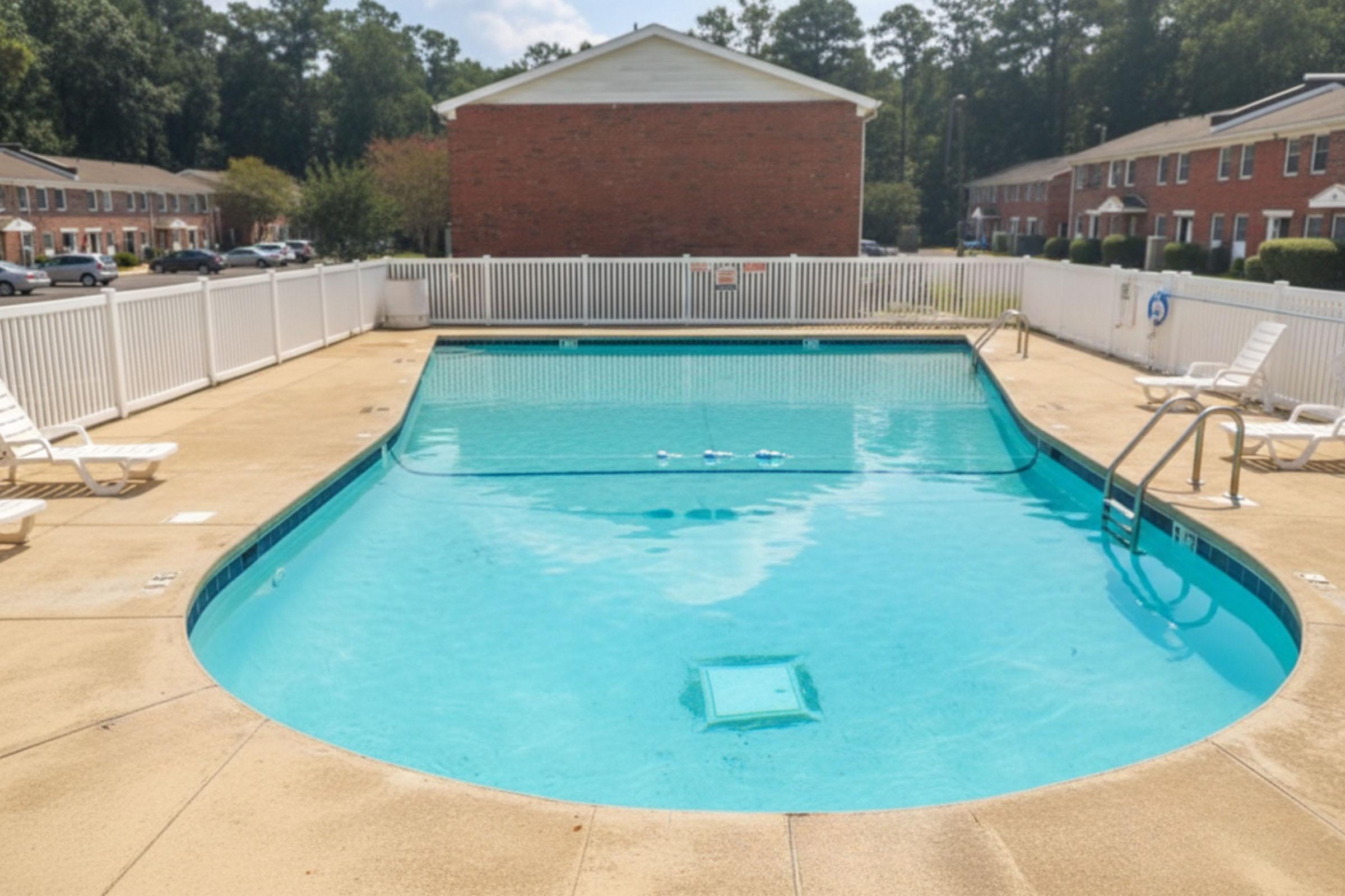 A clear blue swimming pool surrounded by a white fence and lounge chairs, with a few apartment buildings in the background. The pool area is well-maintained and features a diving block, with the water reflecting sunlight.