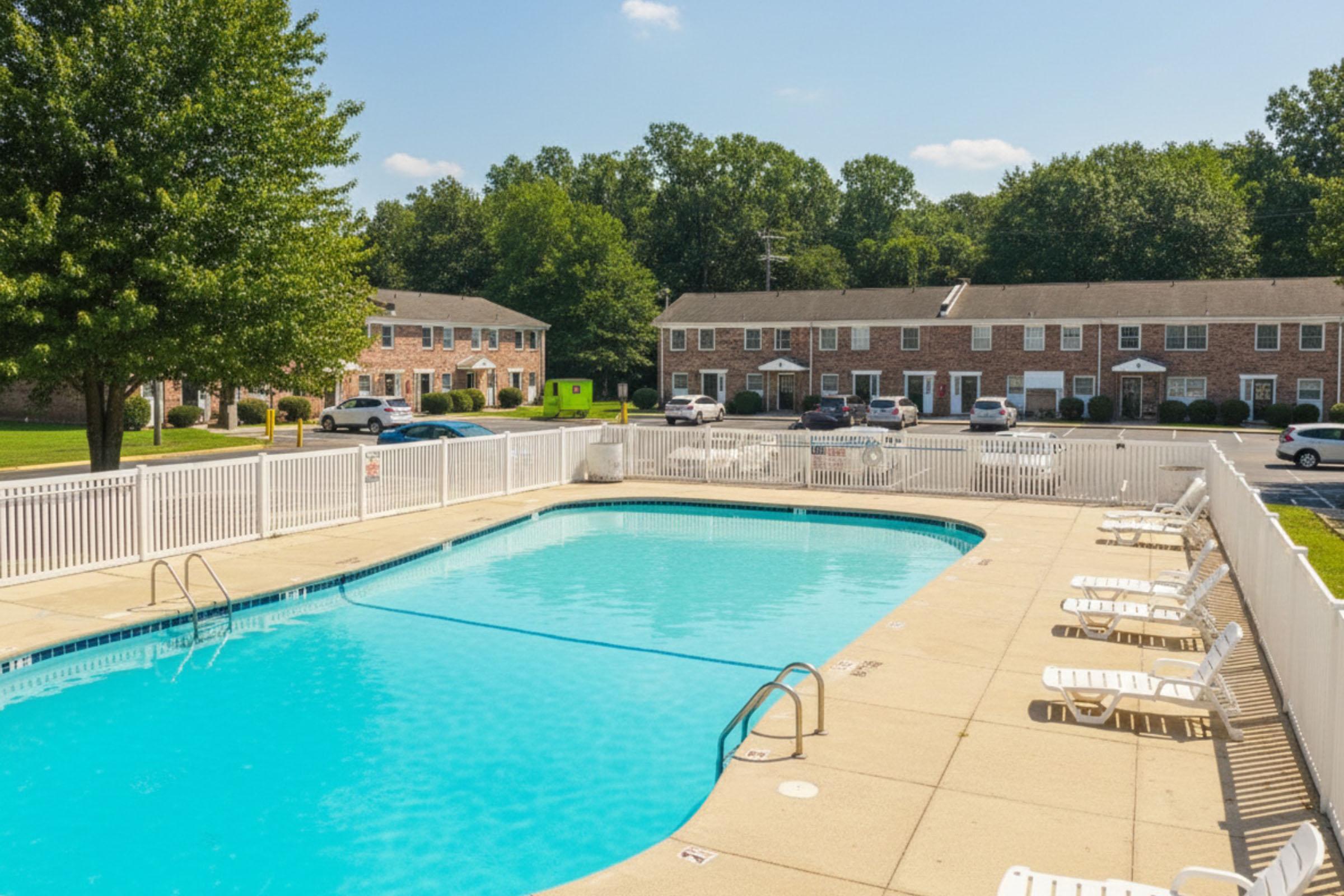 A swimming pool surrounded by a white fence, with lounge chairs positioned around the deck. In the background, there are several brick apartment buildings and green trees under a clear blue sky. The area appears well-maintained and inviting for recreation.