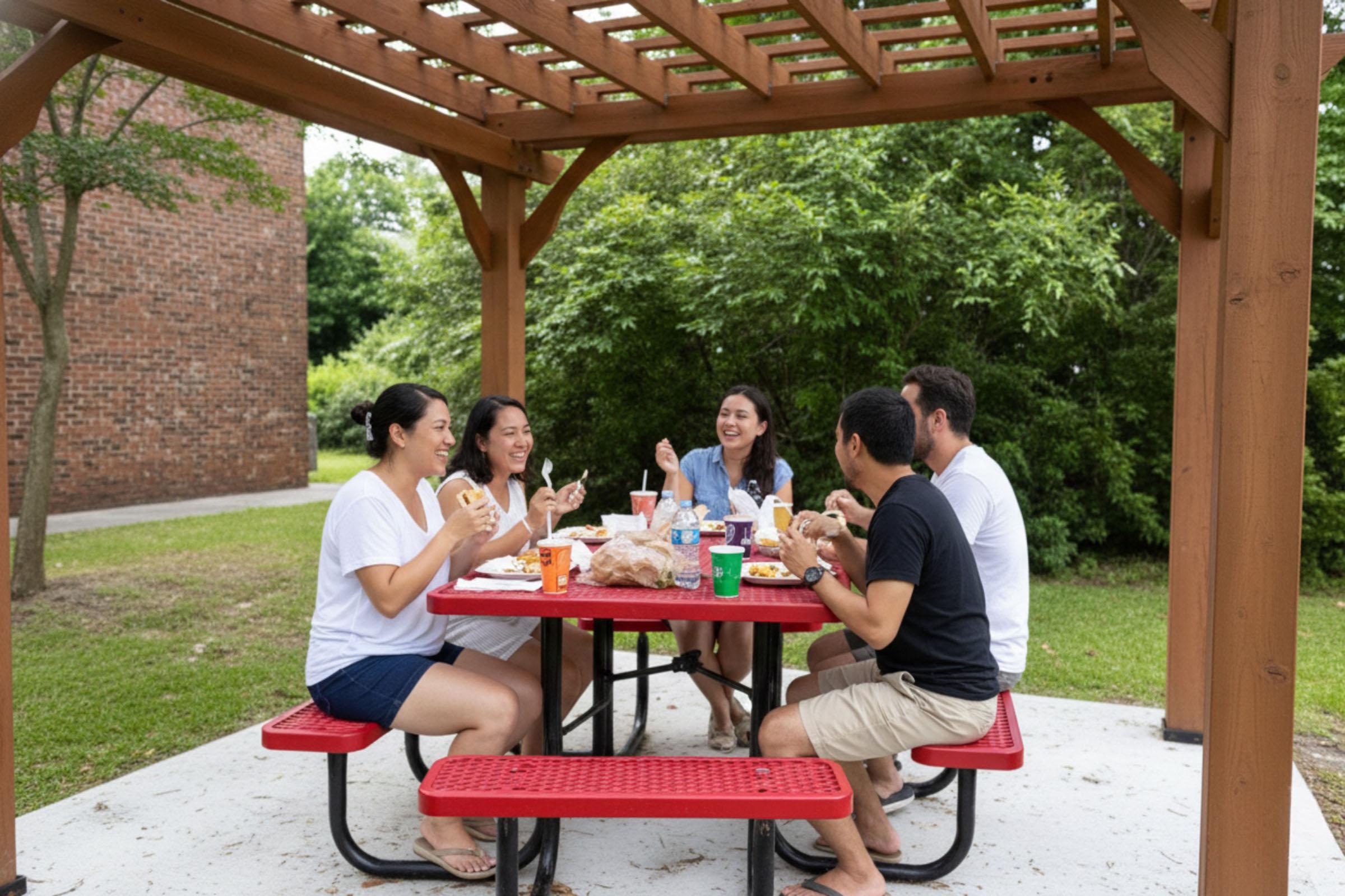 A group of five people enjoying a meal together under a wooden gazebo. They are seated around a red picnic table, sharing food and drinks, and appear to be having a fun, relaxed time outdoors in a green setting.