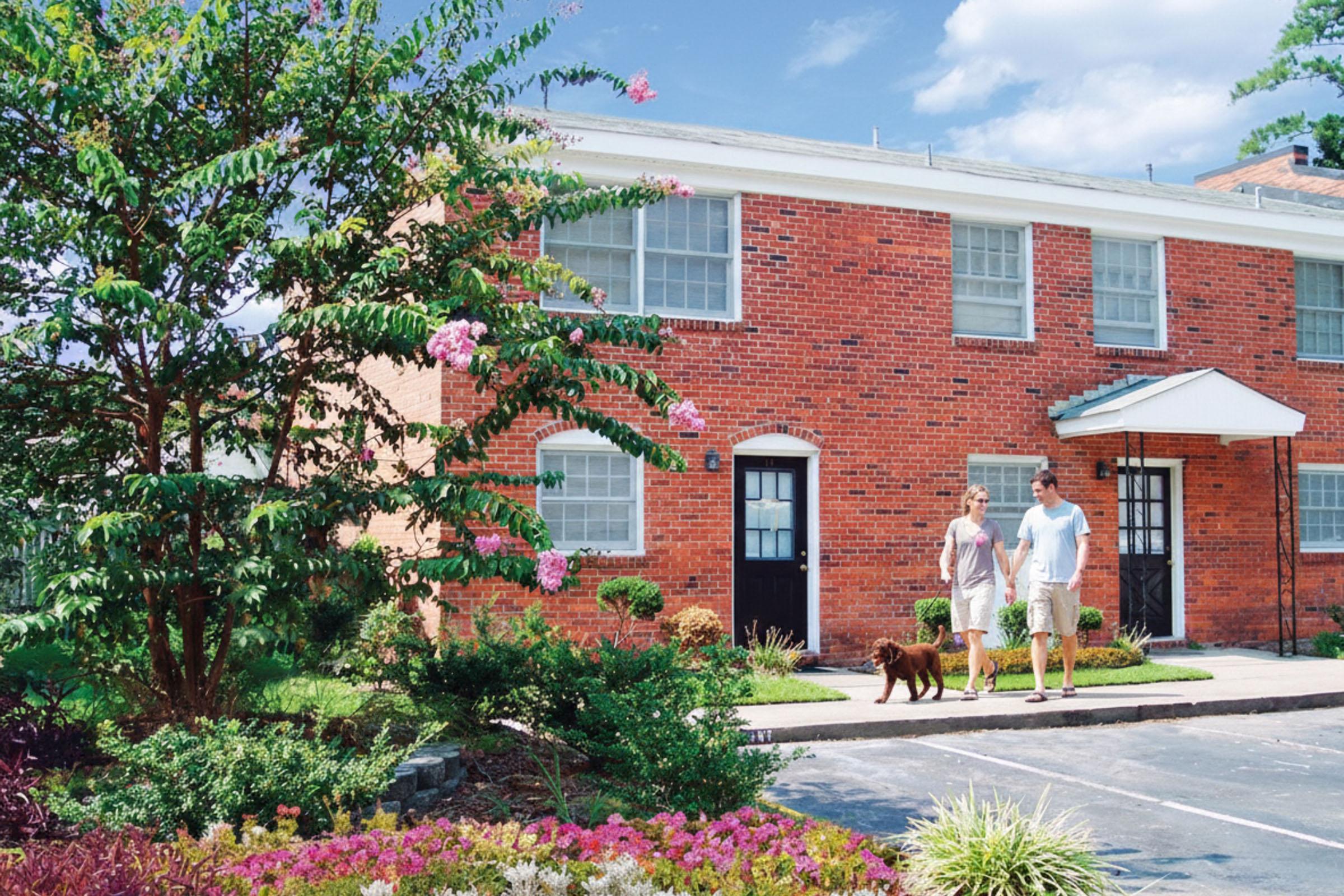 A person and a woman walk a brown dog outside a brick apartment building. The scene features vibrant greenery, colorful flowers, and a clear blue sky, creating a pleasant outdoor atmosphere. The building has multiple windows and a white roof, adding to the appealing residential environment.
