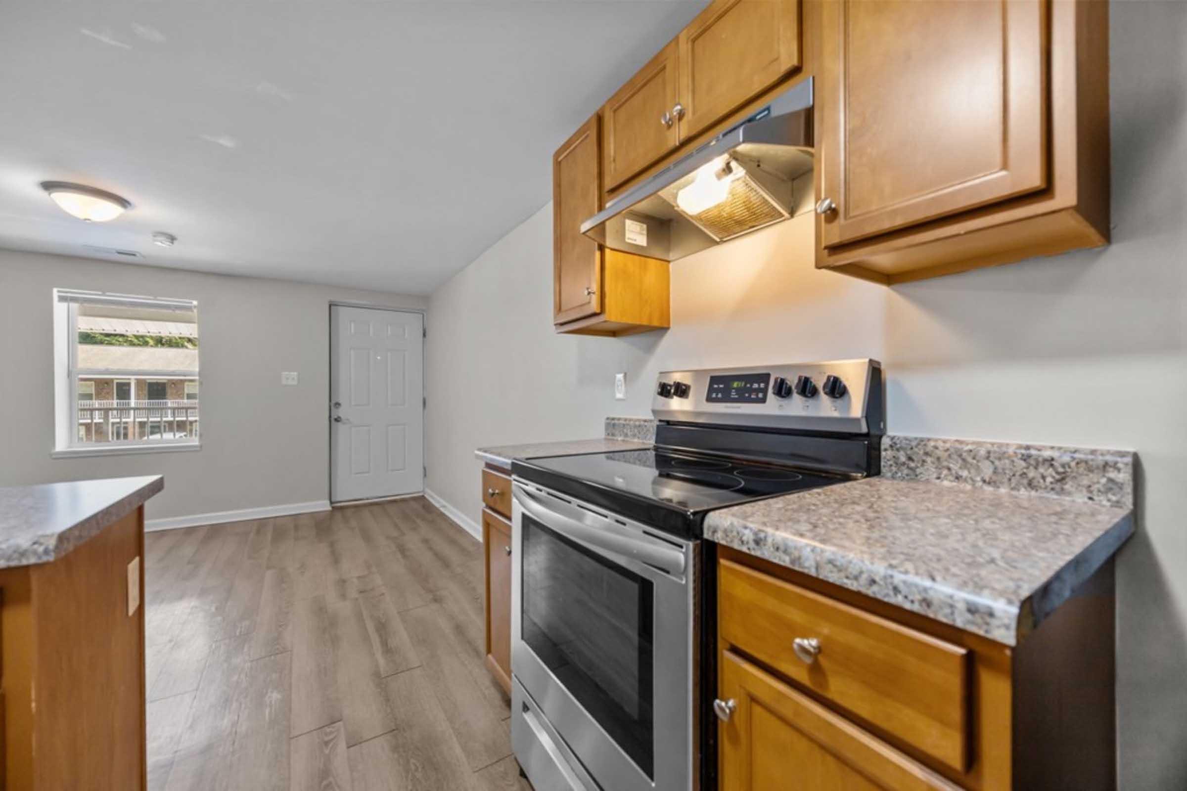 Interior view of a modern kitchen featuring wooden cabinets, a stainless steel stove and oven, and granite countertops. The space is bright, with light coming from a window, and a door visible in the background leading to another area. The flooring is a light-colored wood laminate.