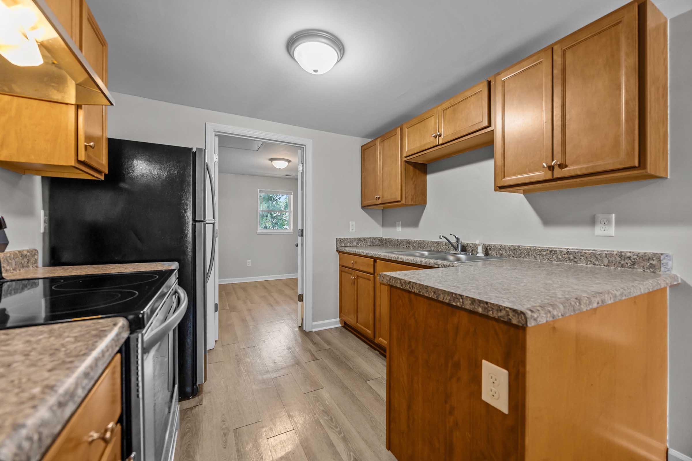 Modern kitchen featuring wooden cabinets, stainless steel appliances, and granite countertops. The layout includes a black refrigerator and stove, with a window allowing natural light. A doorway leads to a adjacent room, enhancing the space. Floor is light-colored wood laminate.