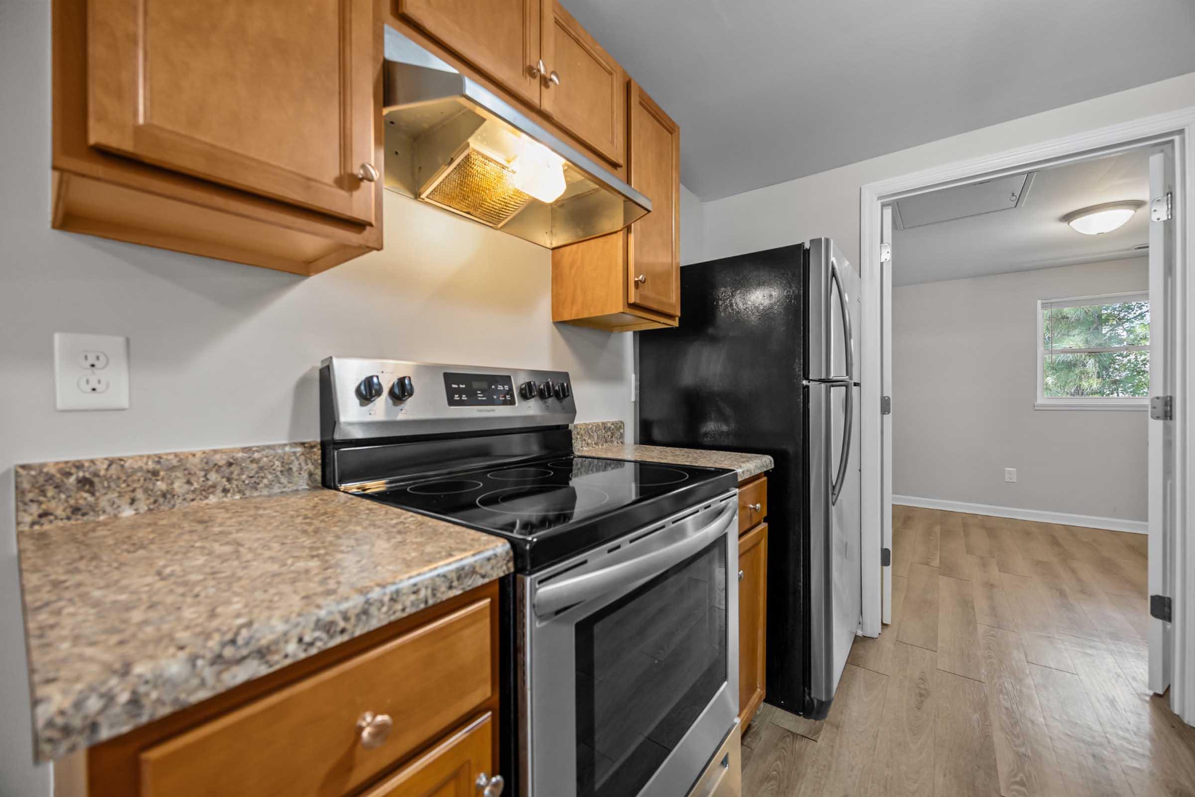 A modern kitchen featuring wooden cabinets, a stainless steel stove, and a black refrigerator. The countertop is made of speckled granite, and there's a door leading to another room, suggesting an open layout. The flooring is light-colored wood, enhancing the overall brightness of the space.
