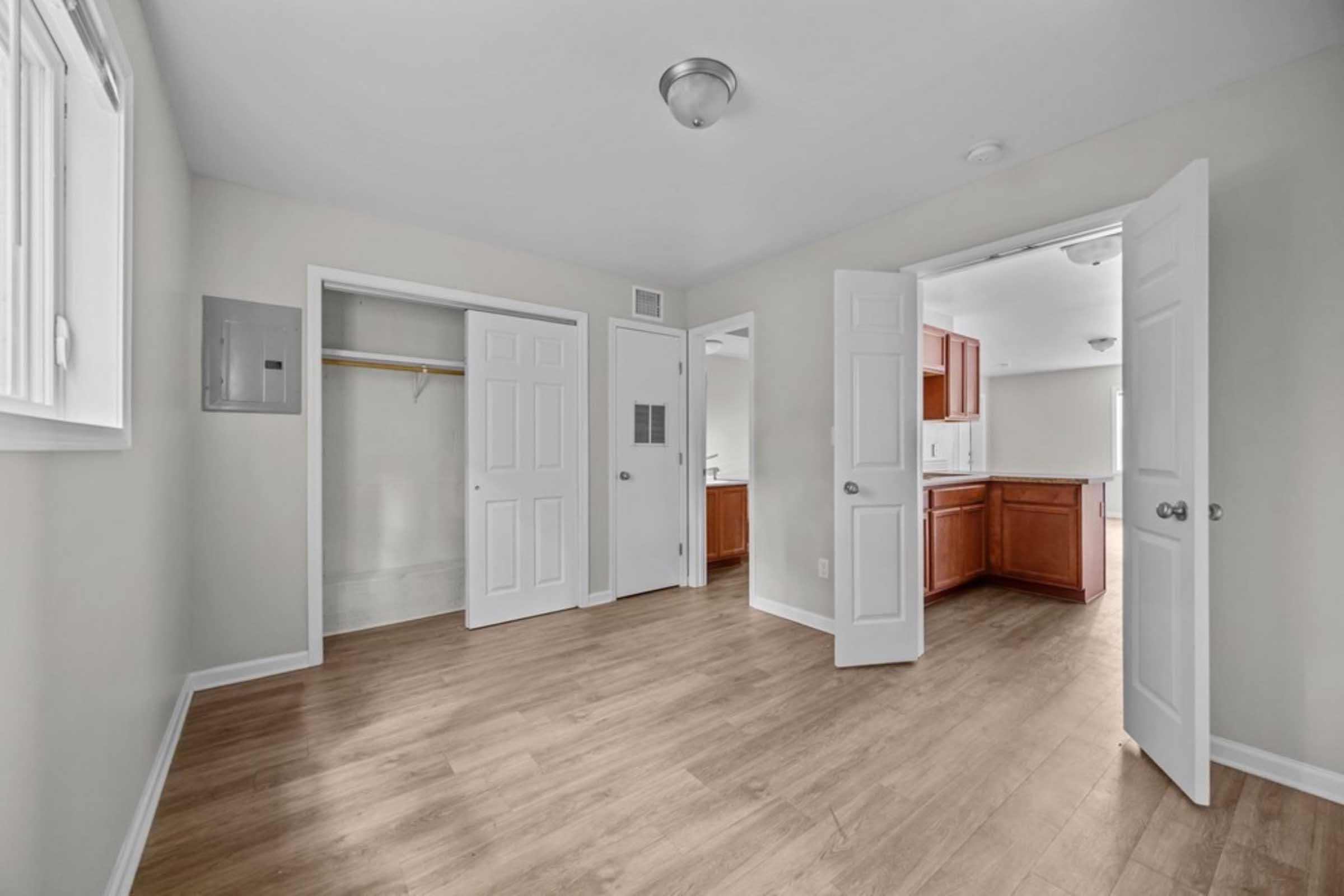 A spacious, well-lit room featuring light wood flooring, white walls, and multiple doors leading to adjacent areas. One door opens to a closet, while another leads to a kitchen space with wooden cabinets visible in the background. The room has a ceiling light fixture and large windows allowing natural light.