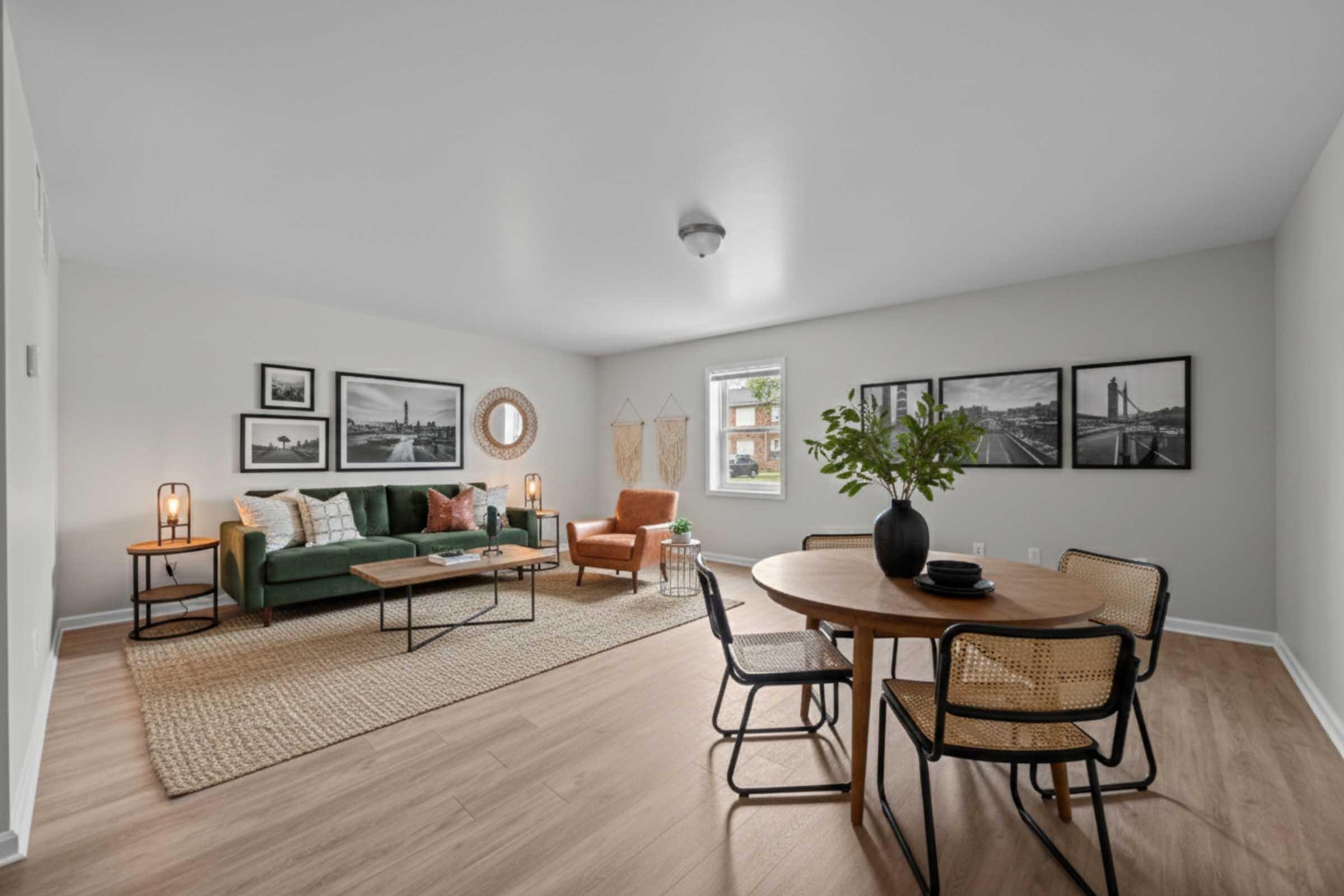 A modern living room featuring a green couch, an orange armchair, and a round wooden dining table. The walls are decorated with black and white photographs, and there's a large round mirror. The floor is light wood, and a textured area rug adds warmth to the space. Plants and stylish lighting enhance the inviting atmosphere.