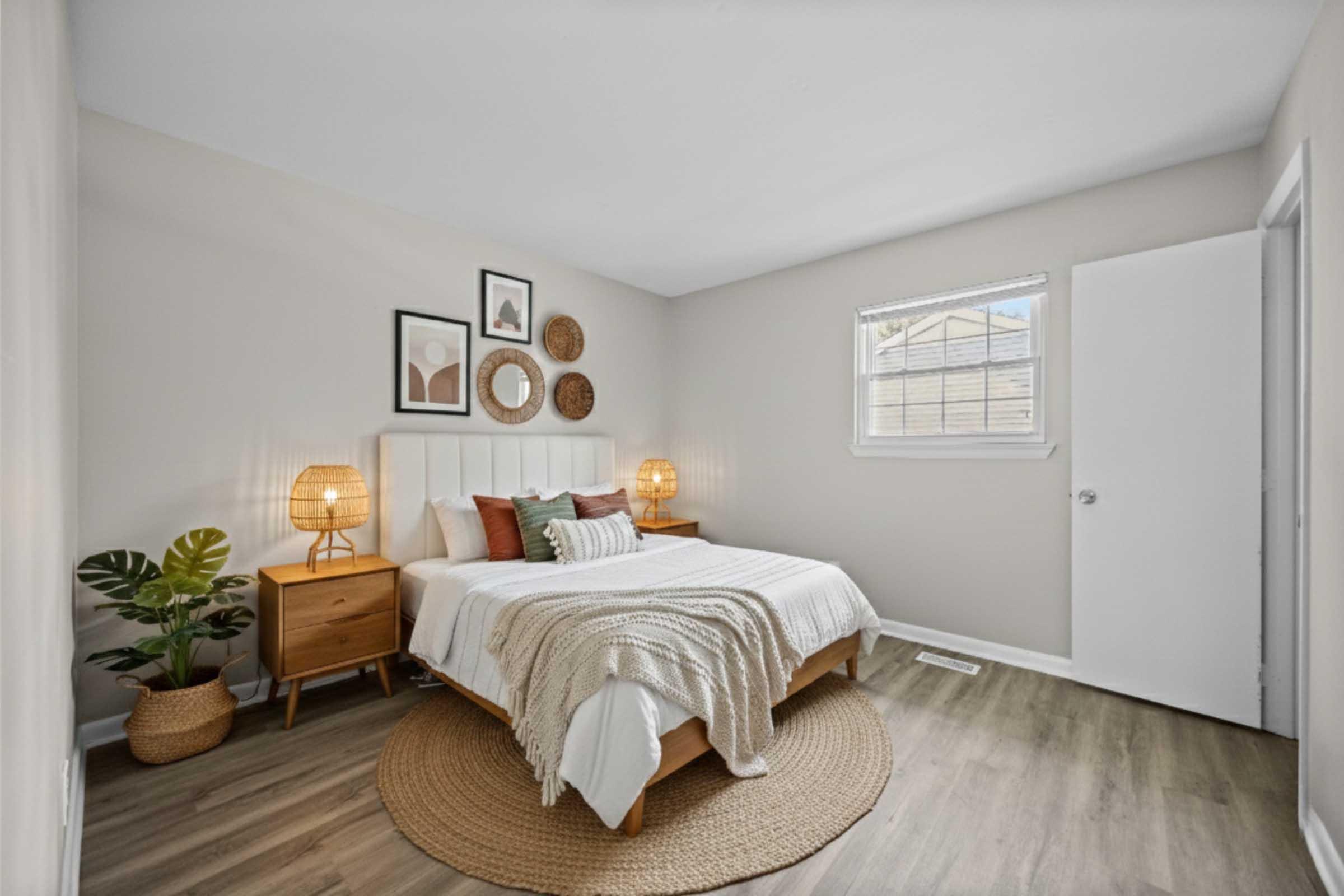 Cozy bedroom featuring a comfortable bed with a white headboard, layered bedding, and decorative pillows. Two bedside lamps illuminate wooden nightstands. Walls are adorned with framed art and woven decor. A plant adds a natural touch, while a round rug complements the space. A window provides natural light.