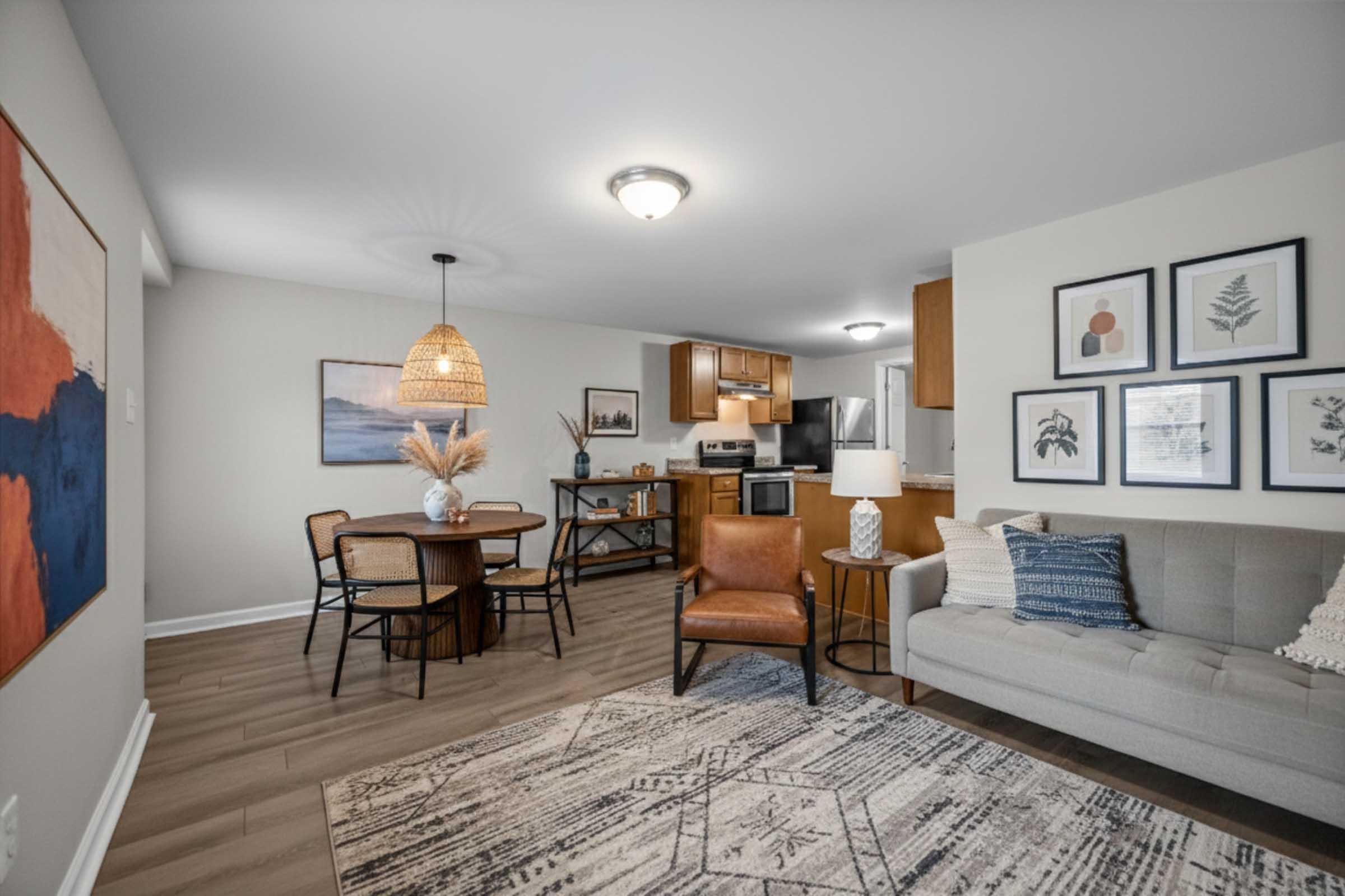 A stylish living area featuring a gray sofa, a wooden dining table with chairs, and modern decor. A pendant light hangs above, and framed artwork adorns the walls. The kitchen is visible in the background, showcasing stainless steel appliances. A cozy rug complements the hardwood floor.