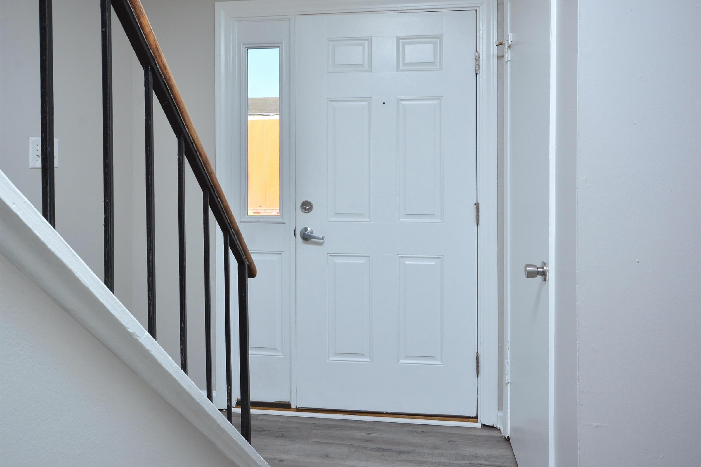 A well-lit entryway featuring a white front door with a window, a staircase with black railings on the left, and light-colored walls. The floor appears to be hardwood or laminate. A small door is partially visible to the right, indicating access to another room.