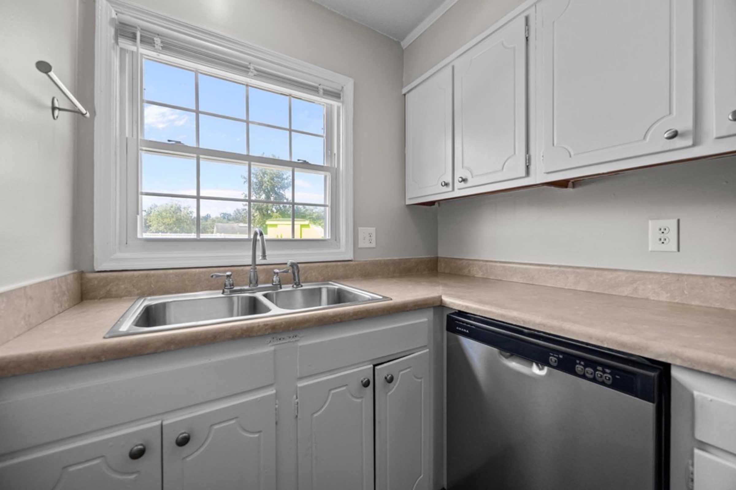 A modern kitchen corner featuring a double sink with two faucets, light-colored countertops, white cabinetry, and a dishwasher. A window above the sink allows natural light to enter, offering a view of the outdoors. The overall design is clean and functional.