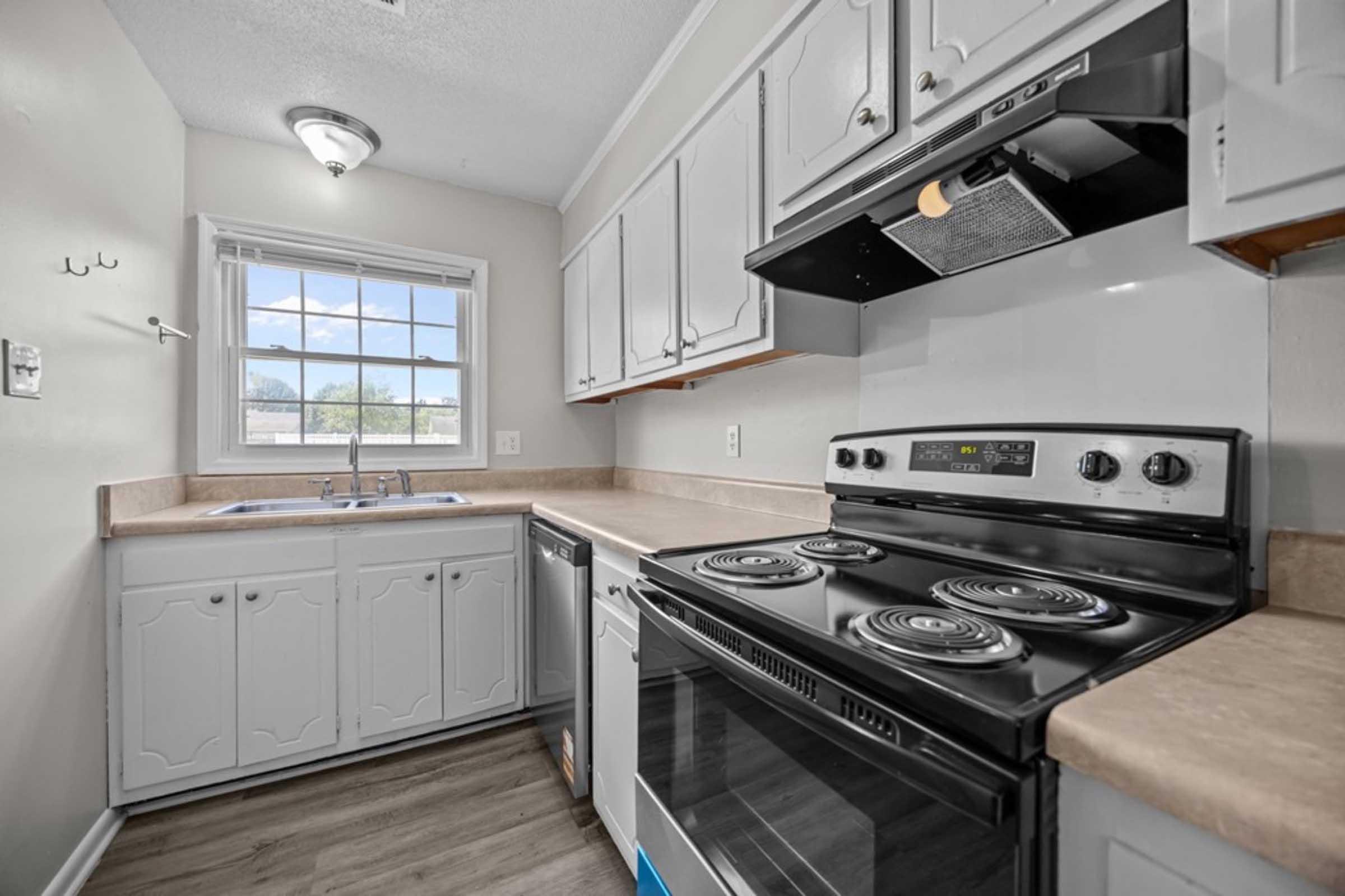 A bright kitchen featuring white cabinetry, a countertop, a black stove with multiple burners, an oven, a dishwasher, and a double sink under a window. The room has light-colored walls and a ceiling light fixture, creating a clean and airy atmosphere.