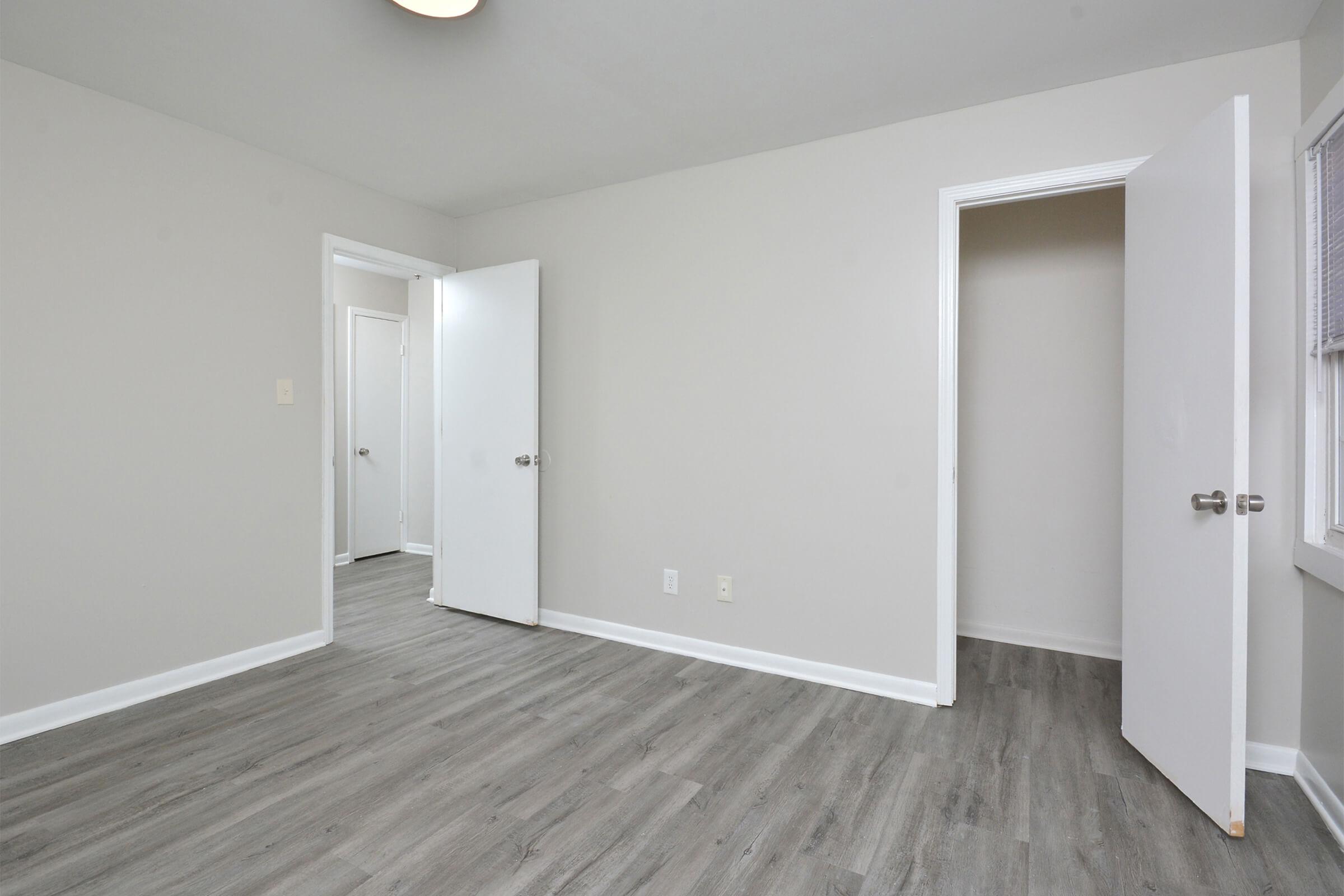 A minimalistic interior room with gray walls and light wooden flooring. Two white doors are visible, one partially open leading to another room. The space is bright and uncluttered, suitable for various uses such as a bedroom or office. A ceiling light fixture is also visible.