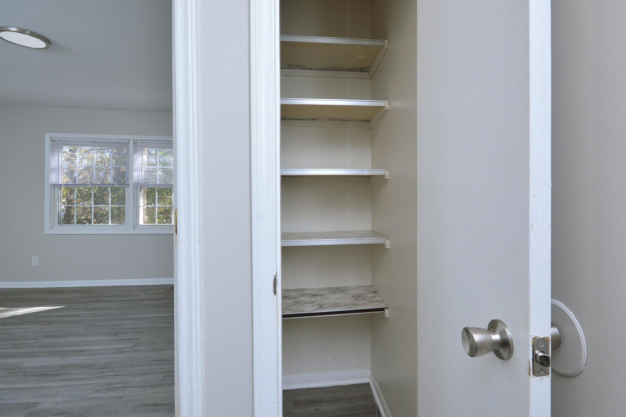 A view into a small closet or pantry with several metal shelves, showcasing a clean, organized space. The area outside features a well-lit room with large windows and gray flooring.