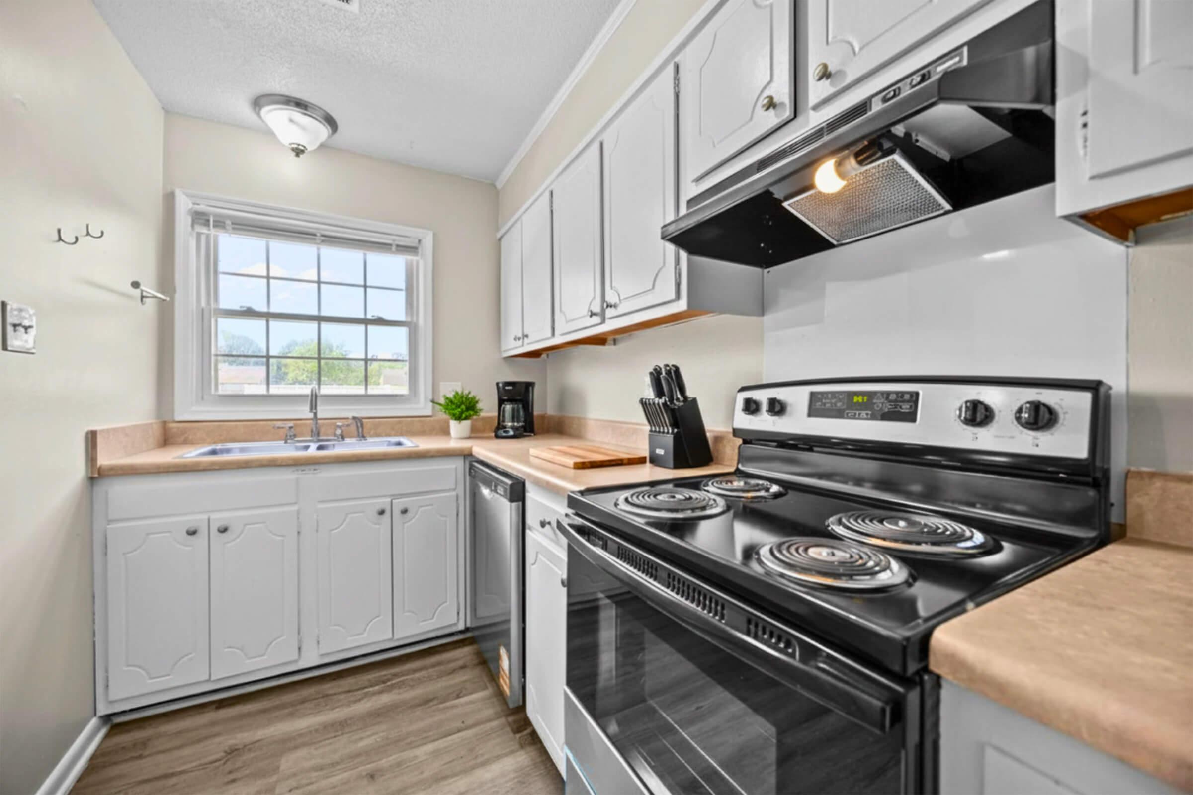 A compact kitchen featuring white cabinets, a window with natural light, a stainless steel stove with an oven, and a microwave. The countertop includes a coffee maker and a knife holder. The space has wood-look flooring and a clean, modern design, emphasizing functionality and bright ambiance.