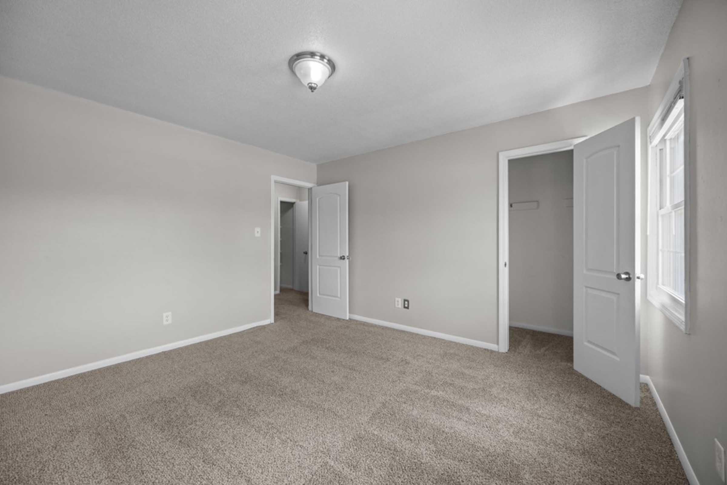 A vacant room with beige walls and carpeted flooring. Two doorways lead to adjacent spaces, with one door slightly open. A ceiling light fixture provides illumination, and a window allows natural light to enter the room.