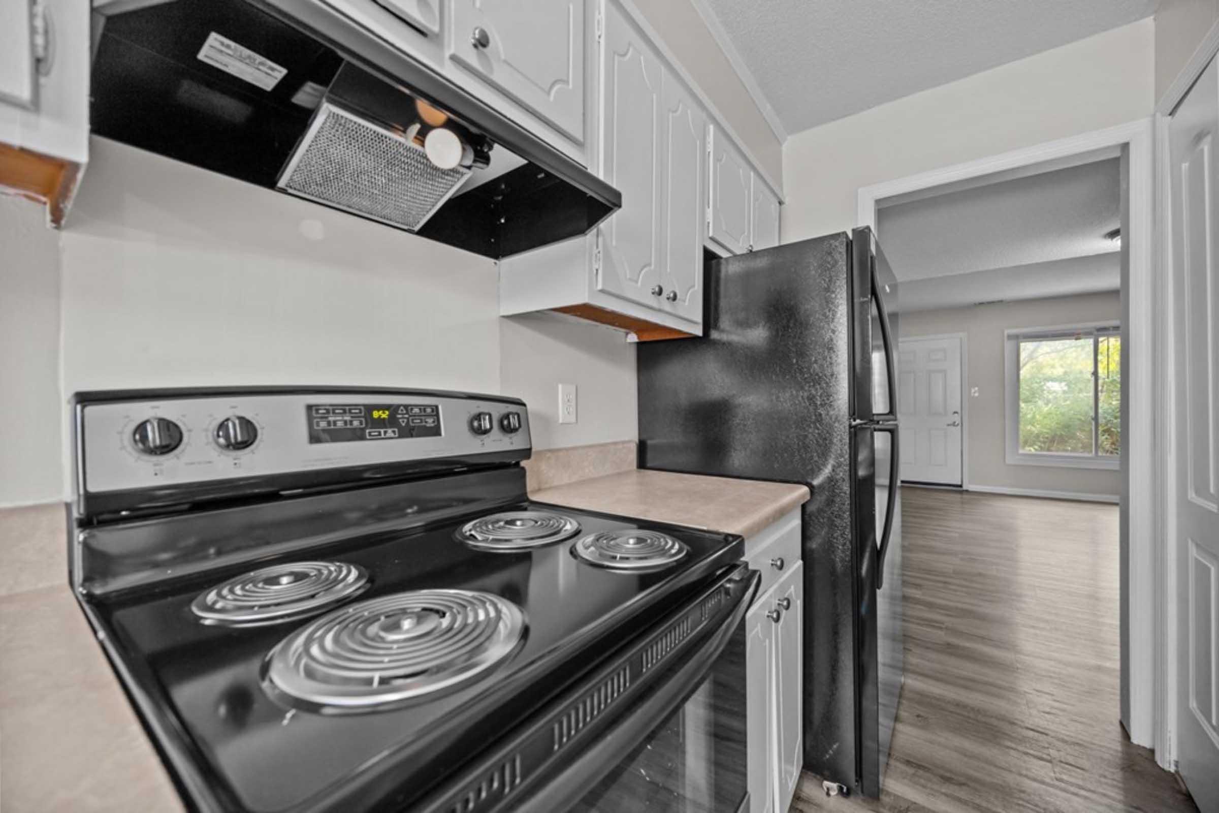 A kitchen featuring a black stove with four burners, overhead cabinets, a hood vent, and a black refrigerator. The room has light-colored walls and a countertop, with an adjacent room visible through an open doorway, showcasing a window with natural light.