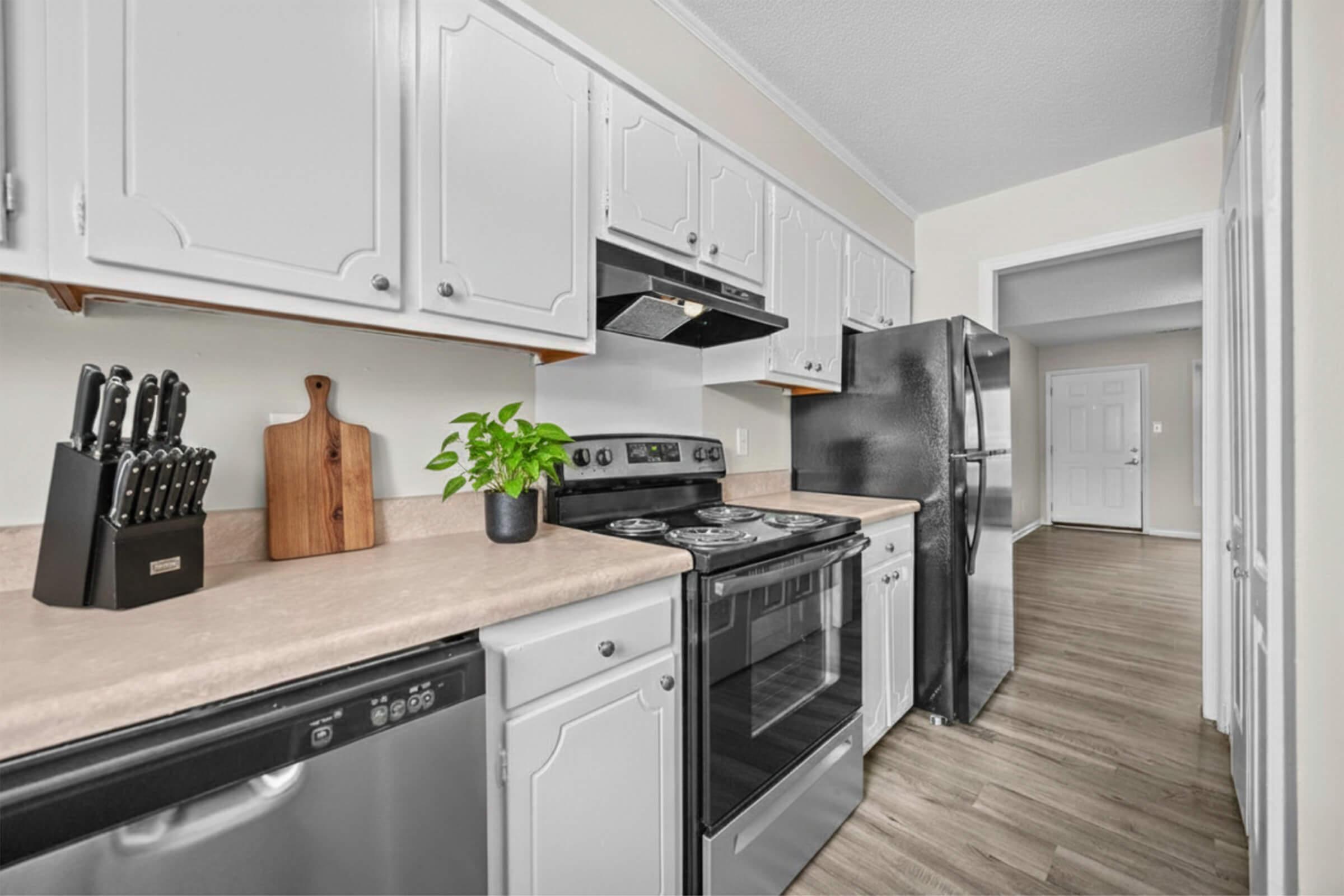A modern kitchen featuring white cabinets, a black refrigerator, and a stainless steel dishwasher. A countertop with a cutting board and a knife set is shown, along with a stove and oven. The flooring is a light wood finish, and a doorway leads to another room.