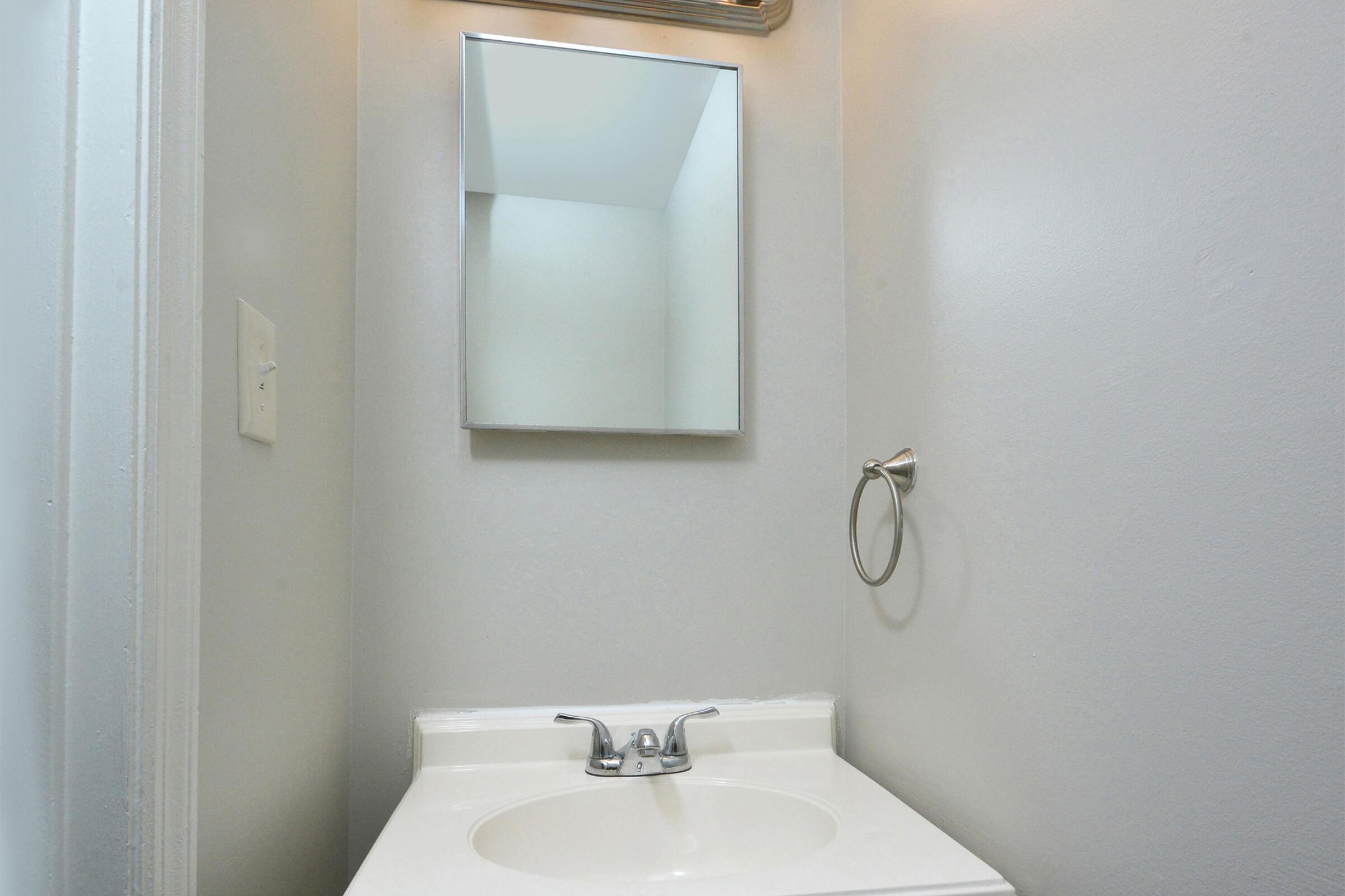 A bathroom sink area featuring a white countertop with a faucet, a rectangular mirror mounted on the wall, and a towel ring. The walls are painted in a light gray color, creating a simple and clean aesthetic.