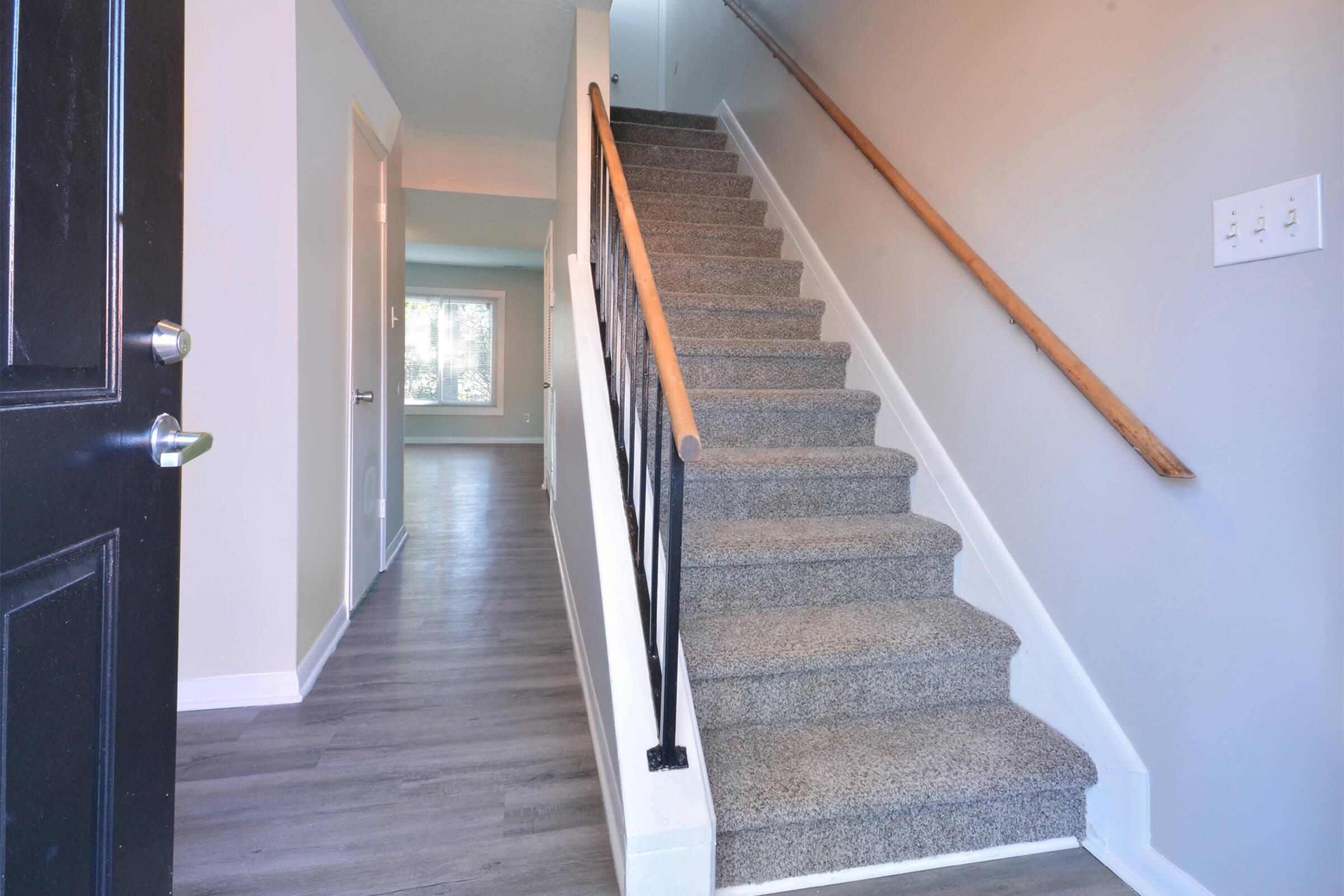 A view of a well-lit entryway featuring a staircase with a wooden handrail, leading to the upper level. The walls are painted in neutral colors, and the flooring is a light-colored laminate. A door is partially visible on the left side, and a window allows natural light into the space.