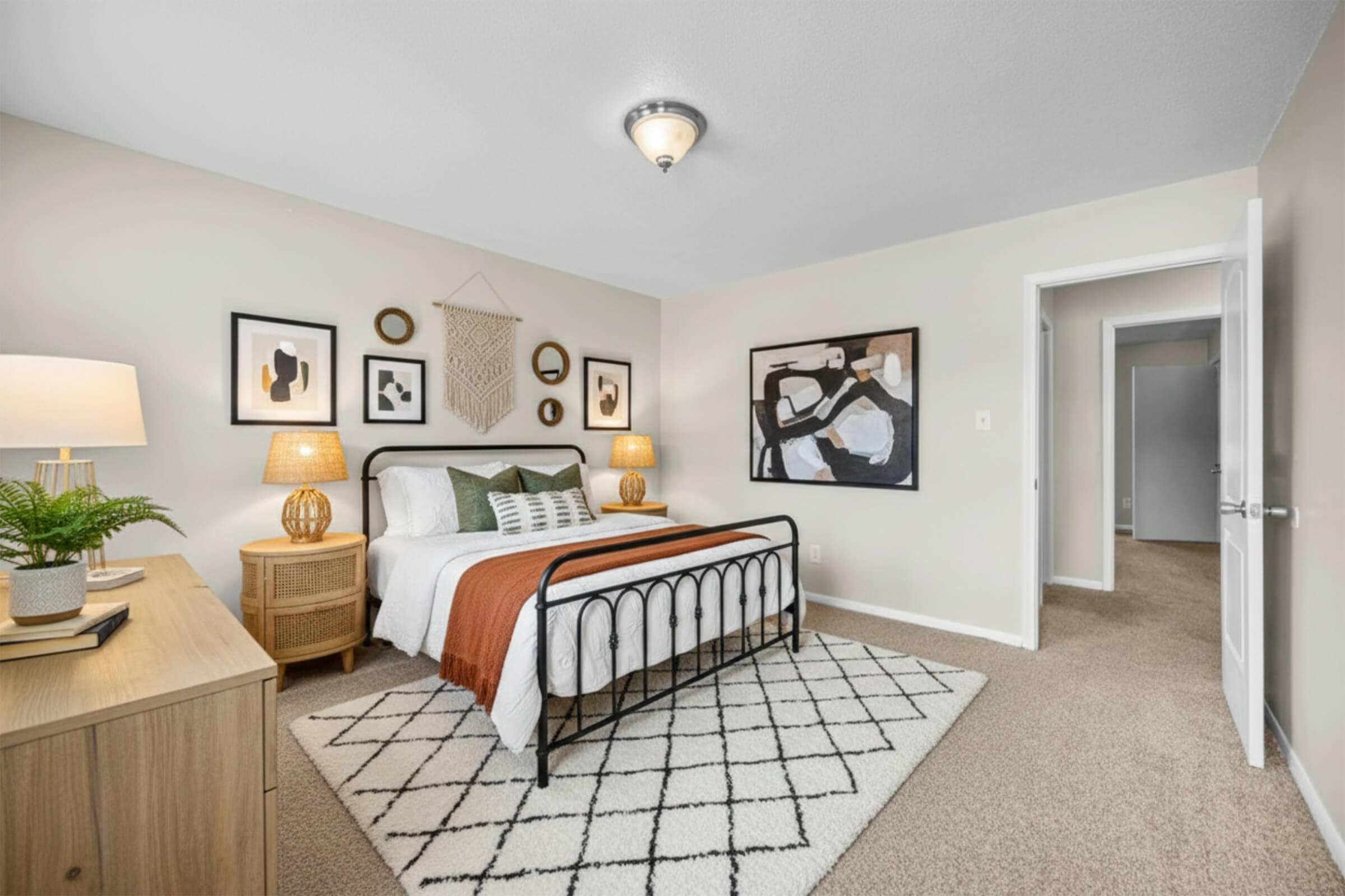 A cozy bedroom featuring a black metal bed frame with white and orange bedding. There's a wooden nightstand with a lamp and decorative items. The walls display framed art pieces, and a woven wall hanging adds texture. A light-colored rug is on the carpeted floor, with a doorway leading to another room.