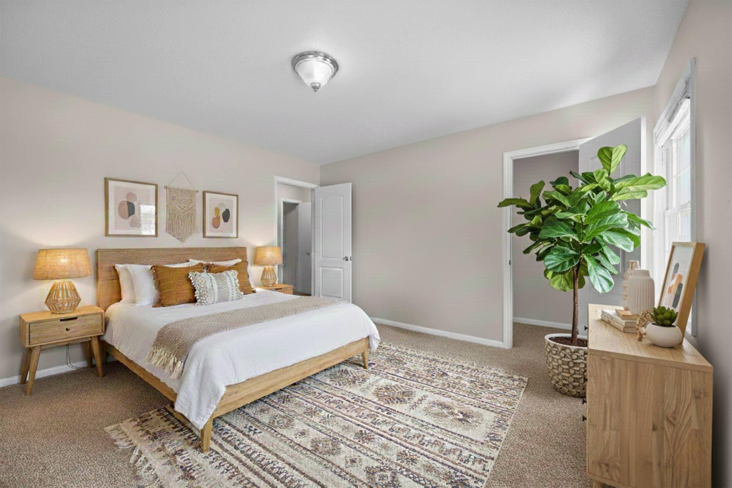 A cozy bedroom featuring a light-colored wall, a wooden bed with decorative pillows, and two bedside lamps. There's a patterned area rug on the floor, a potted plant in the corner, and a wooden dresser with decorative items. A doorway leads to another room, adding to the welcoming atmosphere.