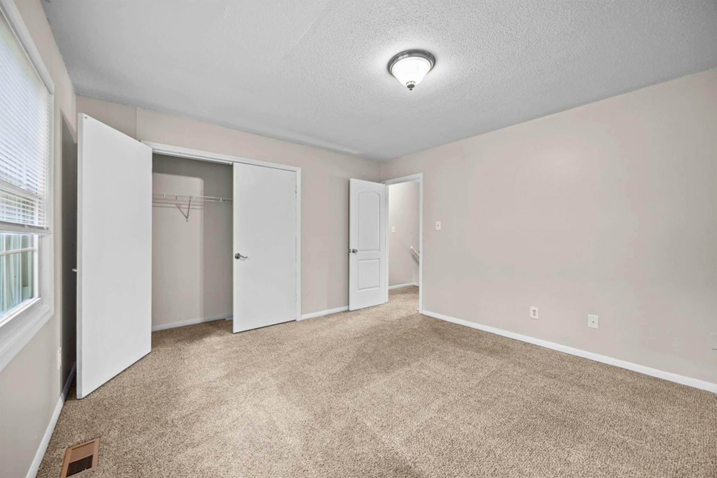 A spacious, empty room with beige walls and carpet. Two open doors lead to a closet and another area. A ceiling light fixture is visible, and a window lets in natural light, highlighting the neutral color scheme. The room is well-lit and ready for furnishing.